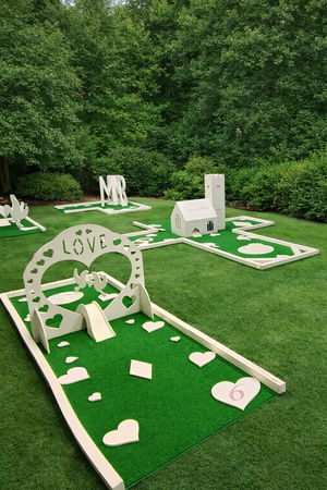 Miniature themed outdoor mini golf course with love and heart decor, small white playhouse, and green grass, surrounded by trees.