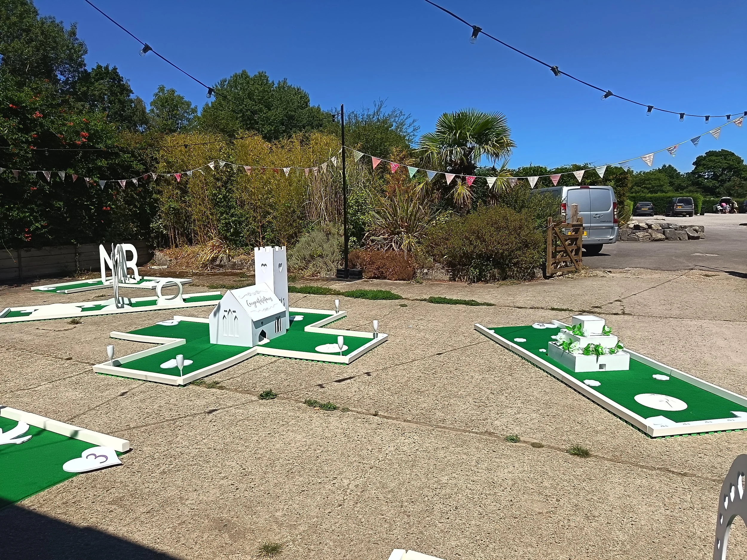 Outdoor mini golf course with green turf, white obstacles, and a small white house decoration, surrounded by trees, string lights, and parked cars under a blue sky.