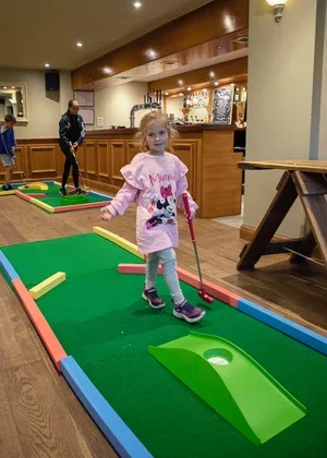 A young girl playing mini golf in an indoor entertainment center, with other children and a wooden bar in the background.