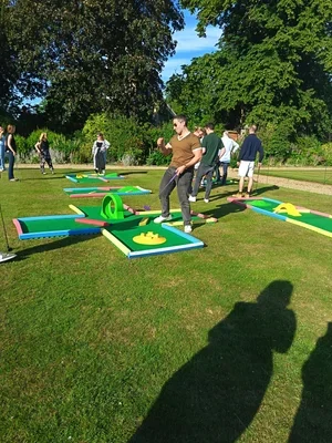 People playing mini-golf on a grassy field with large trees in the background.