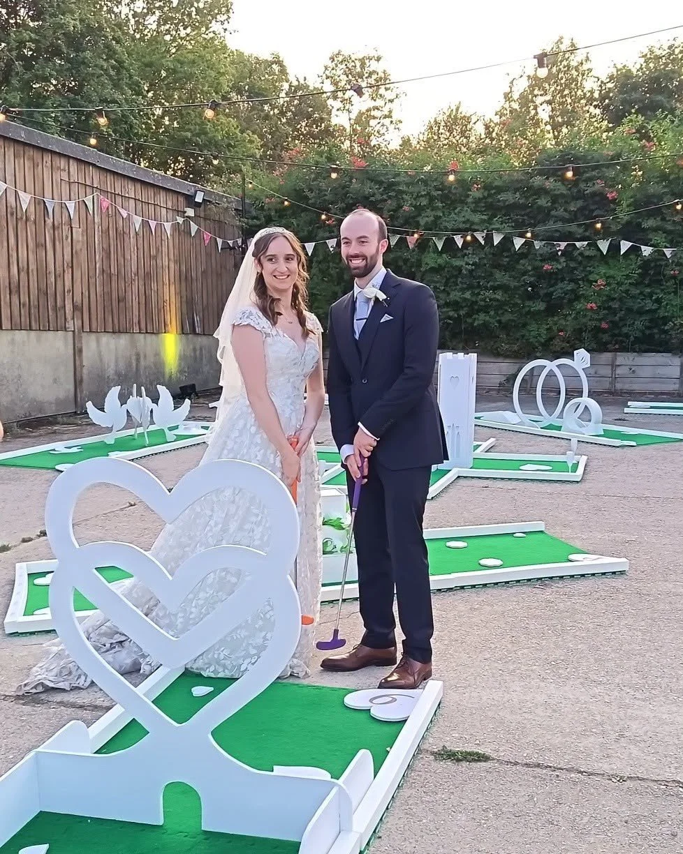 A bride and groom smiling at a wedding reception outdoors, standing on a mini-golf course with decorative white hearts and rings. String lights and bunting decorate the area, with trees and a wooden fence in the background.