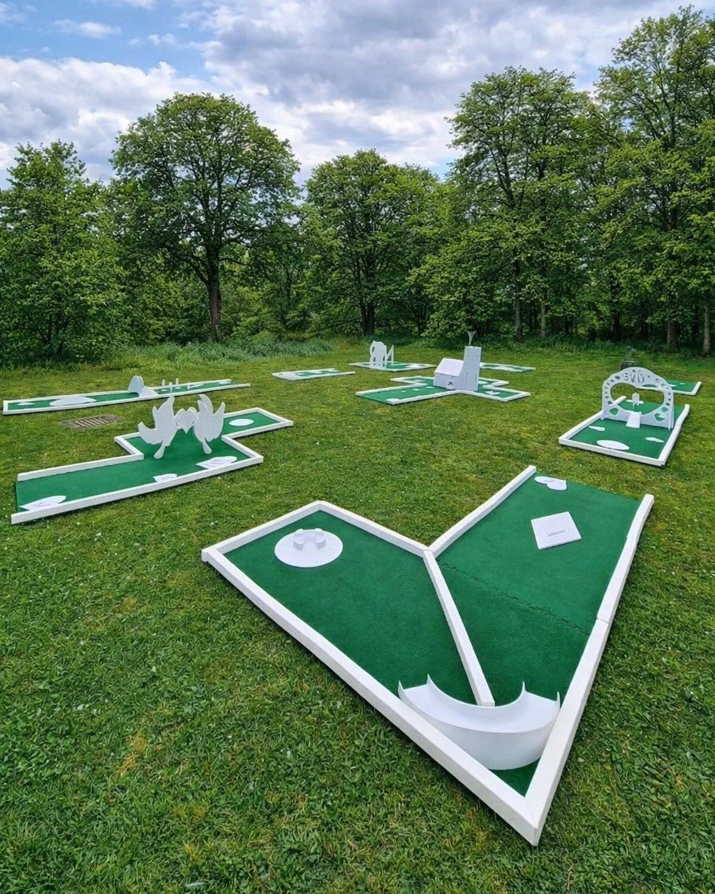 Outdoor mini golf course with several green turf holes and white decorations, surrounded by trees and cloudy sky.