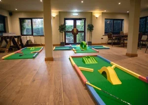 Indoor miniature golf course with multiple holes, green artificial turf, and colorful obstacles, in a well-lit room with large windows and wooden flooring.