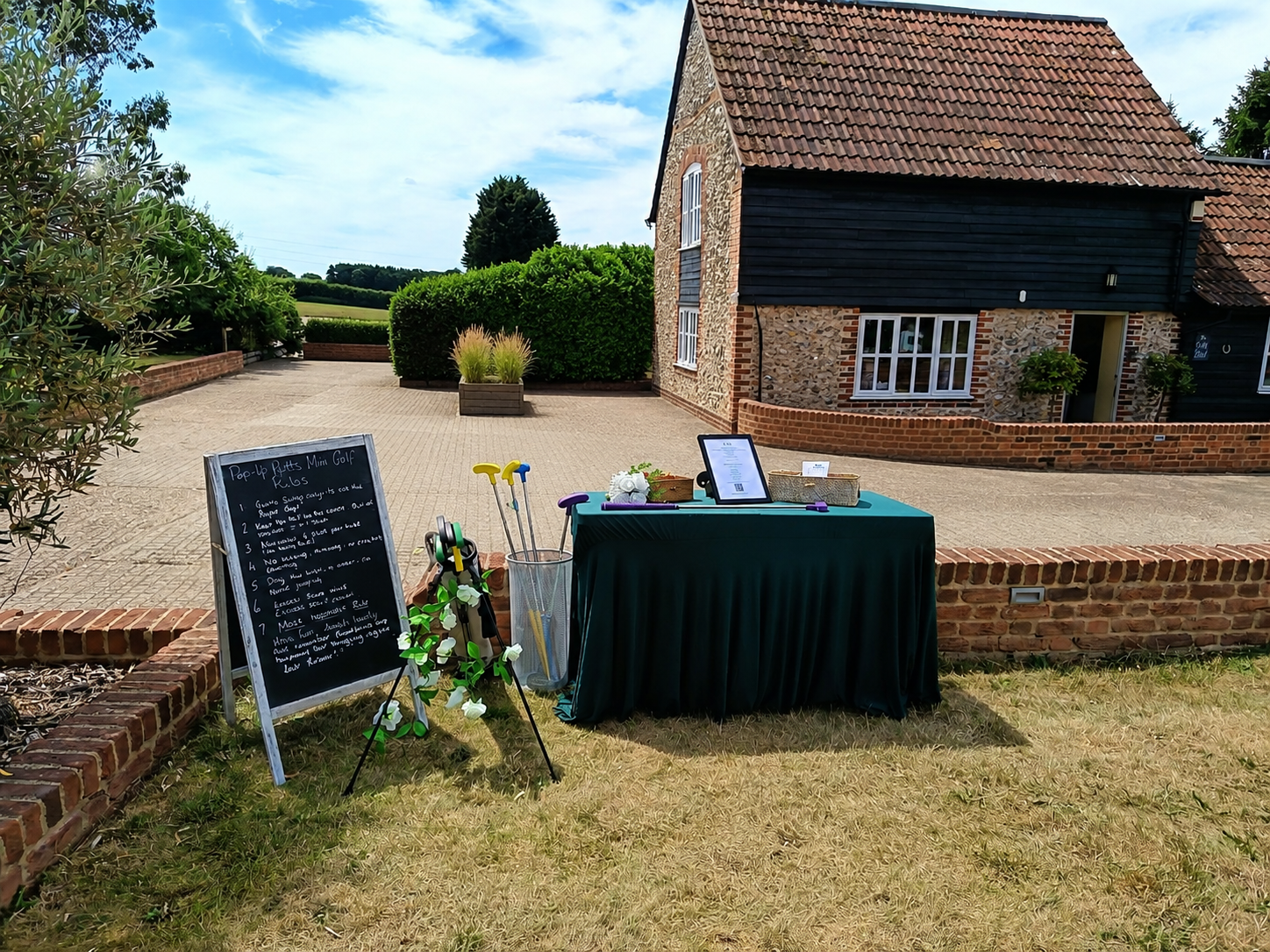 Outdoor scene with a blackboard, golf clubs, and a table with items on it, set up on a lawn in front of a stone and wood house with a tiled roof, under a blue sky with clouds.