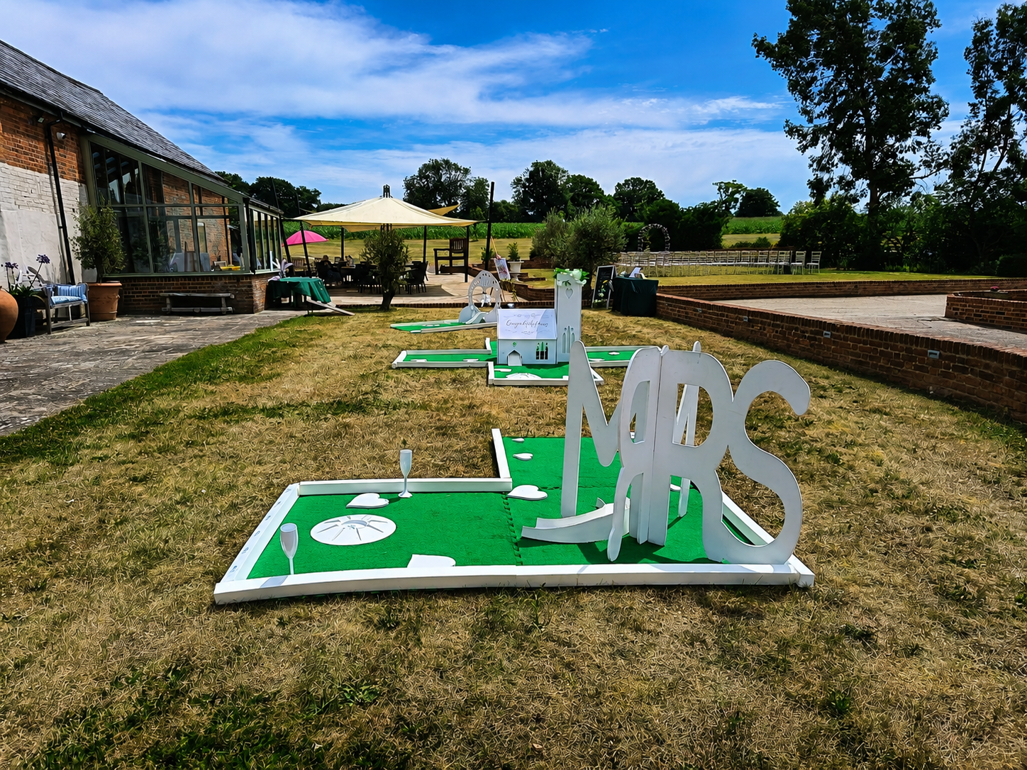 Outdoor miniature golf course set up on a grassy lawn with decorative signs spelling "NAGS" in large white letters, nearby patio area with tables and umbrellas, brick building and trees in the background under a partly cloudy sky.