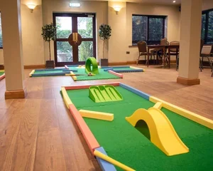 Miniature golf setup with two holes, small ramp, and green turf in an indoor room with chairs and windows