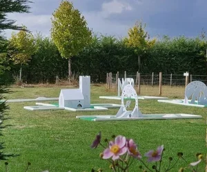 Outdoor scene with various white and gray playground equipment on a grassy lawn, trees in the background, and pink flowers in the foreground.