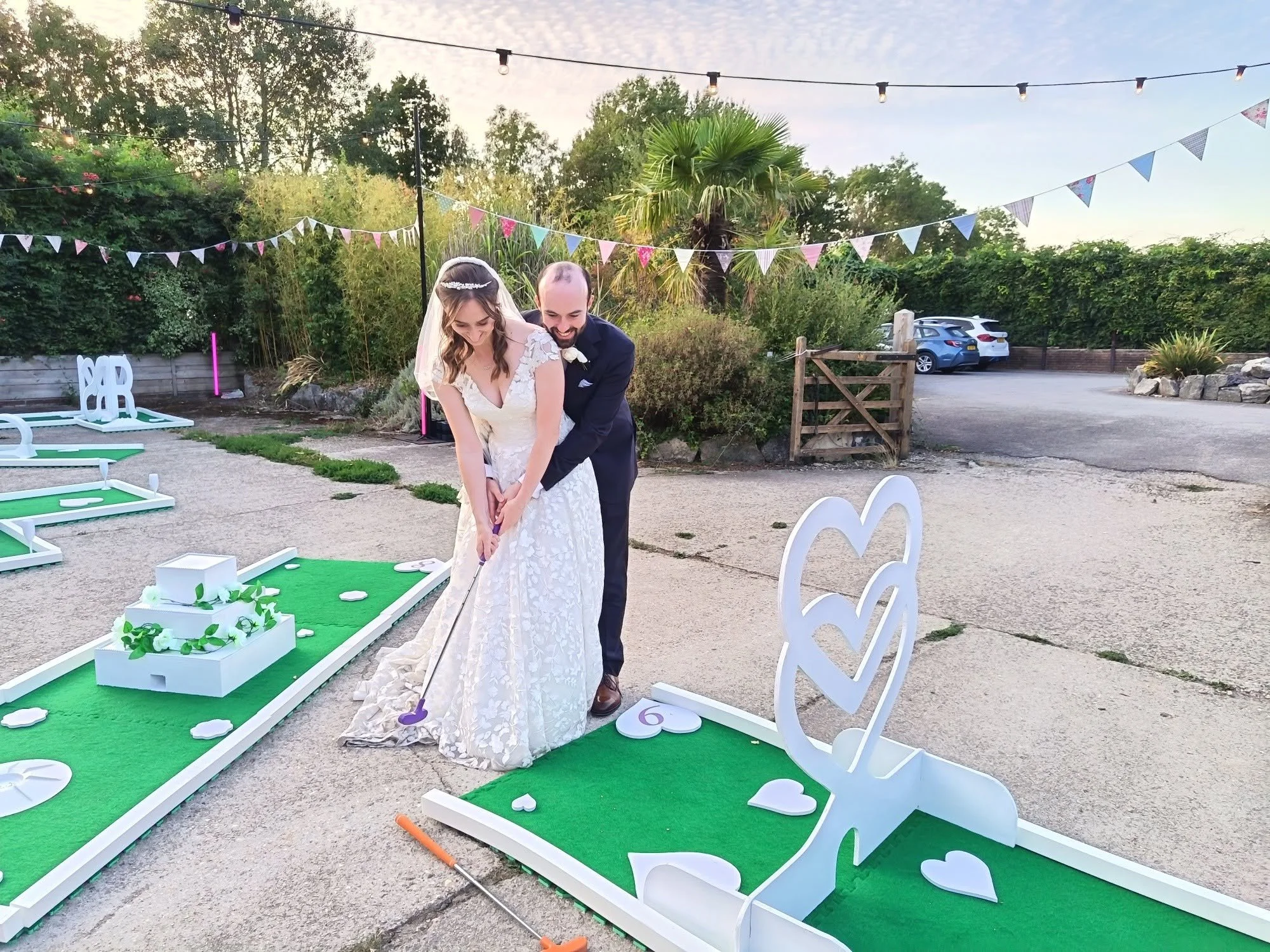 A bride and groom playing mini-golf at an outdoor wedding reception. The bride is in a white wedding dress with lace details, and the groom is in a dark suit. They are smiling and bent over, holding a mini-golf club together. Decorations include pink, blue, and white bunting hanging overhead, and white heart-shaped signs on a small artificial grass course. The background features trees, parked cars, and a wooden gate.