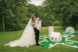 A bride and groom standing on a miniature golf course, with the bride in a wedding dress and veil, and the groom in a white shirt and dark pants, surrounded by trees and grass.