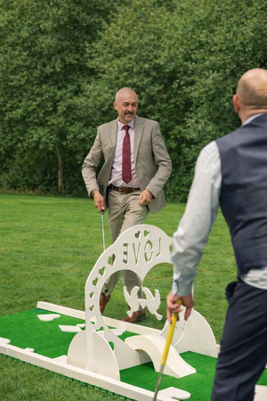 Two men playing miniature golf outdoors on a grassy course with trees in the background. One man, in a beige suit and red tie, is holding a golf club and smiling at the other man, who is in a gray vest and white shirt, holding a golf club. There is a white decorative structure with the word 'LOVE' cut out, serving as a golf obstacle.