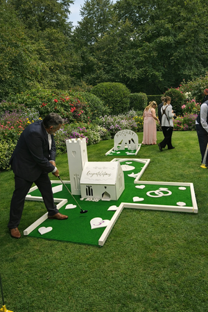 People playing miniature golf in a garden with bushes and flowers, with a white castle-themed structure.
