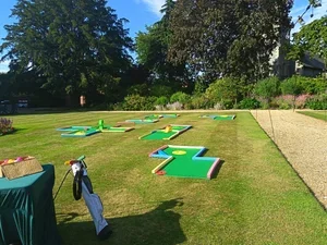 A mini golf course set up outdoors on a grassy area with several colorful holes and putters. There is a green table with golf balls and a bag of golf clubs nearby, trees, and bushes in the background.