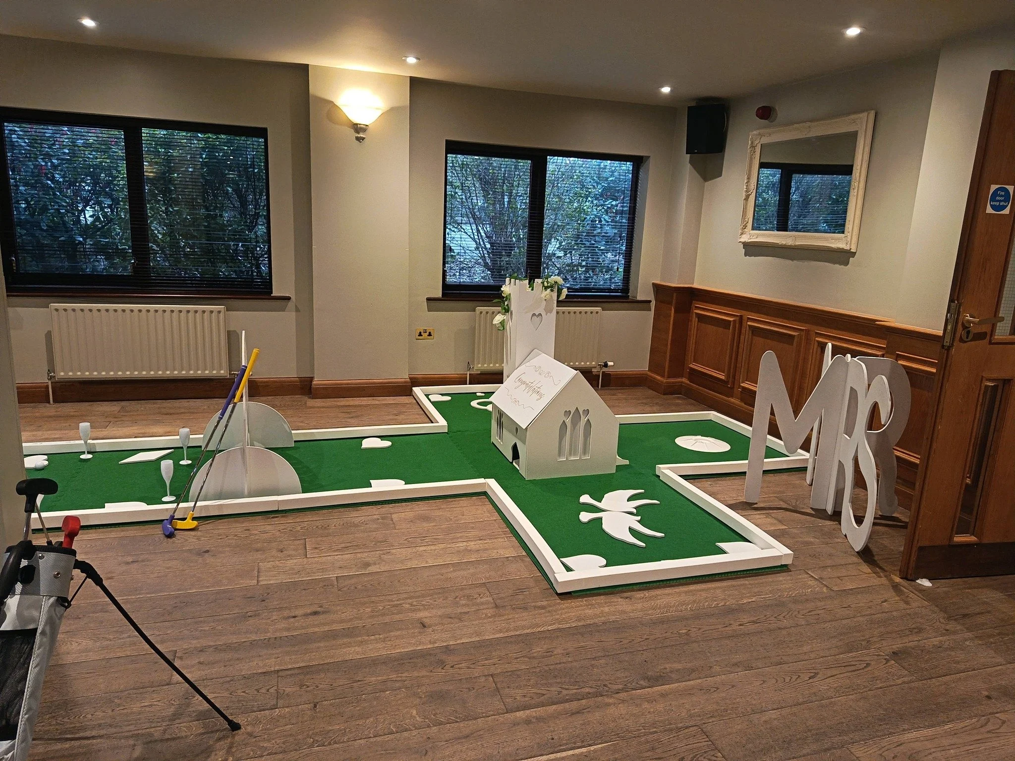 Indoor room with three windows showing trees outside, a wall-mounted lamp, a mirror with ornate white frame, and a wooden paneling on the wall. There is a green miniature golf course setup on the floor with white decorative figures, a small white house, and golf clubs. A sign on the house reads 'Congratulations.'