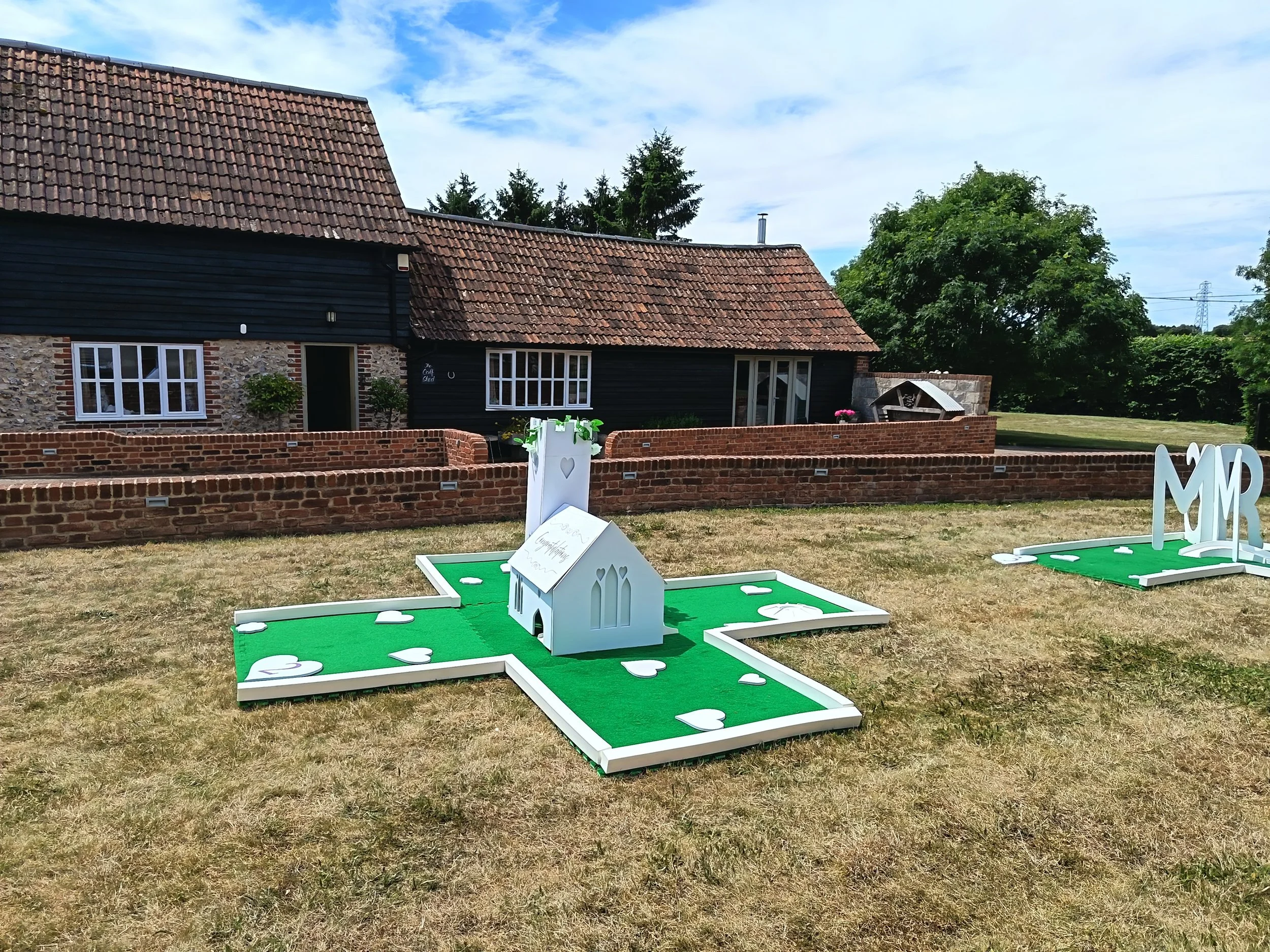 Outdoor scene with a brick building with a tiled roof, a black wooden wall, and several white window frames. Pink flowers, green trees, a lawn, and outdoor games with green mats and white wooden structures are visible in the foreground.