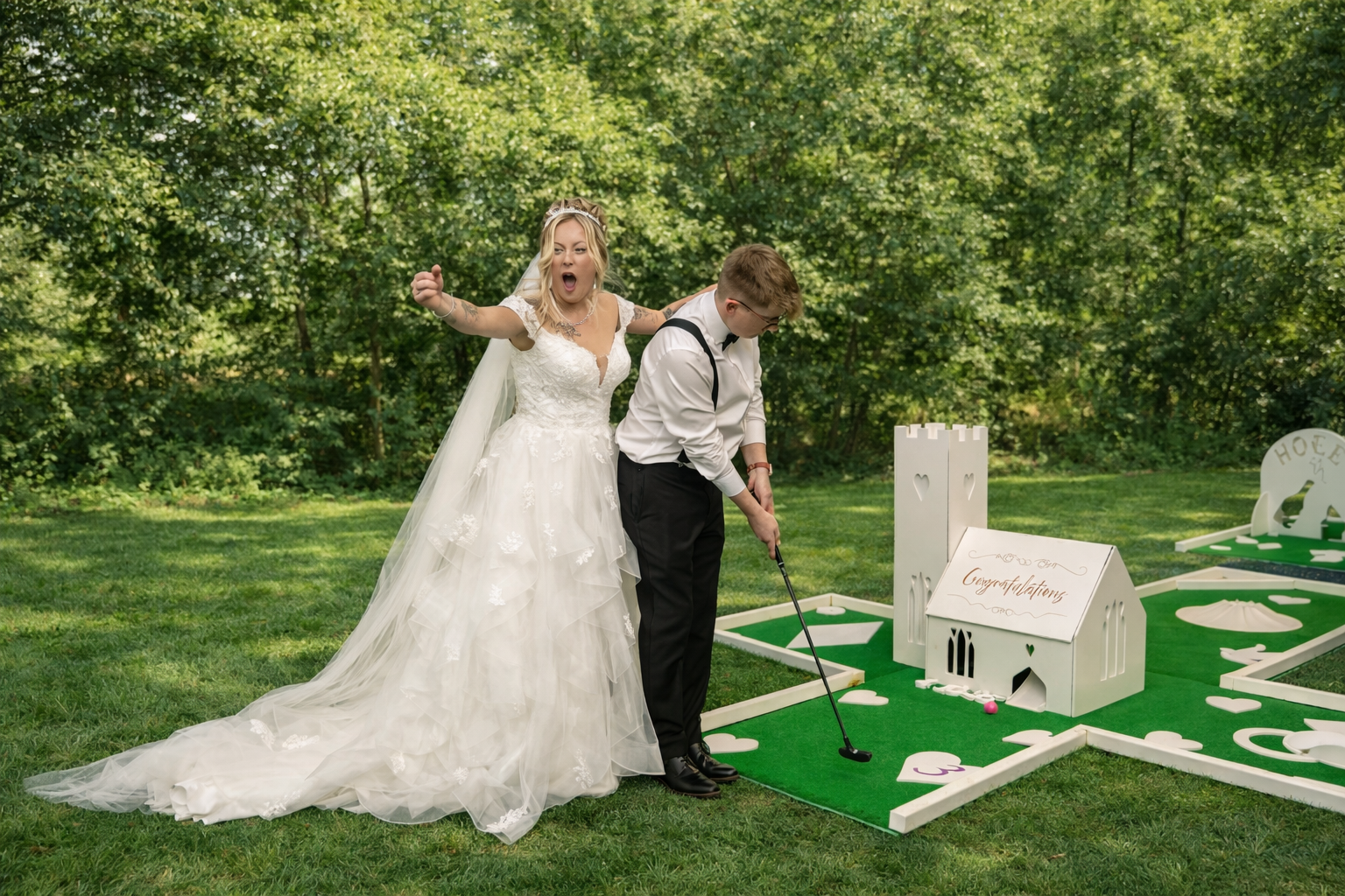 Bride and groom playing mini golf outdoors, with the bride wearing a white wedding dress and the groom in black pants, white shirt, suspenders, and glasses. The bride appears to be reacting excitedly as the groom prepares to putt on a green mini golf course with decorative structures.