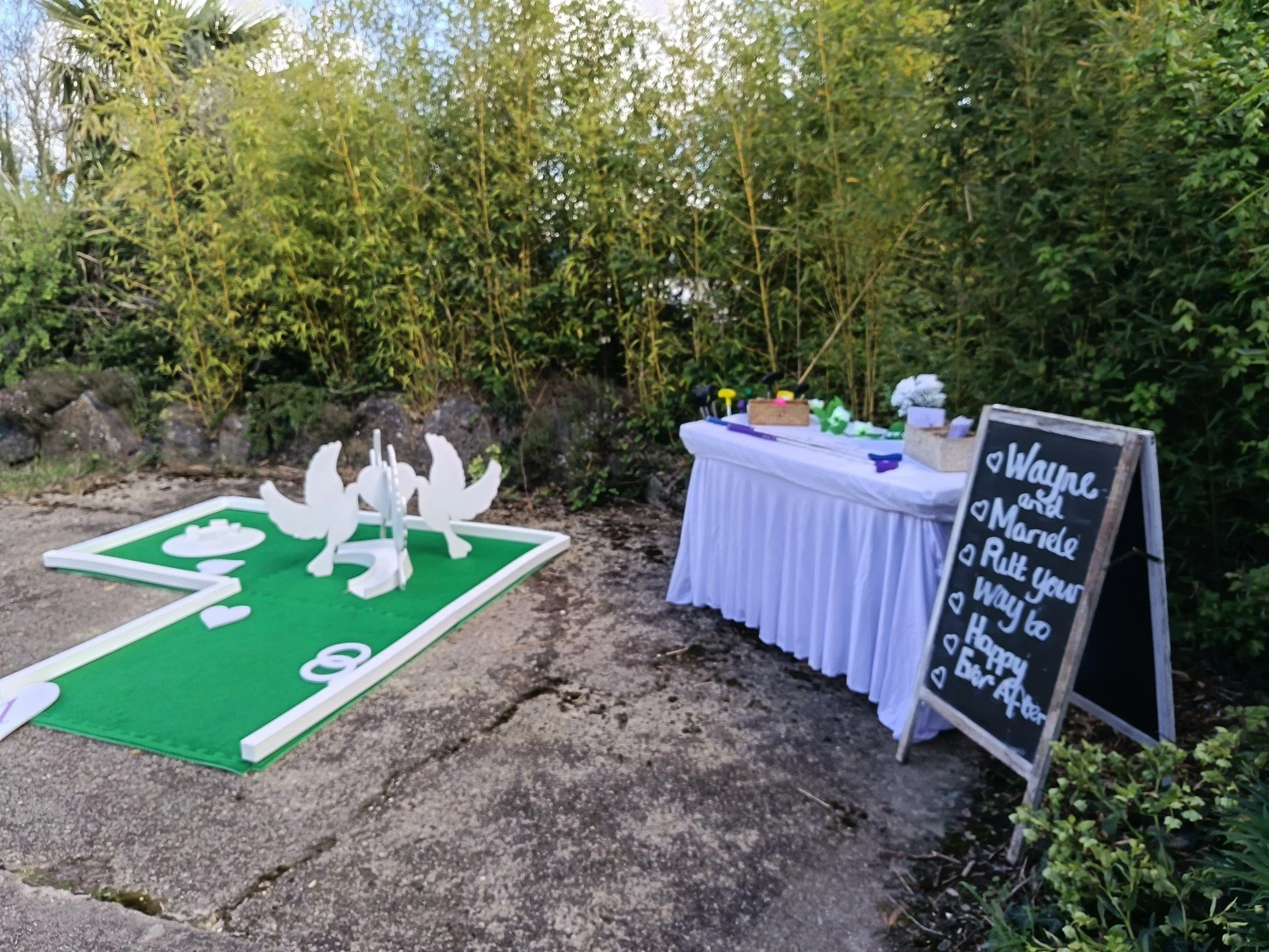 Outdoor yard wedding or celebration setup with a white tablecloth-covered table, decorative blackboard sign with a message, and a cornhole game with white game pieces. Surrounded by greenery and rocks.