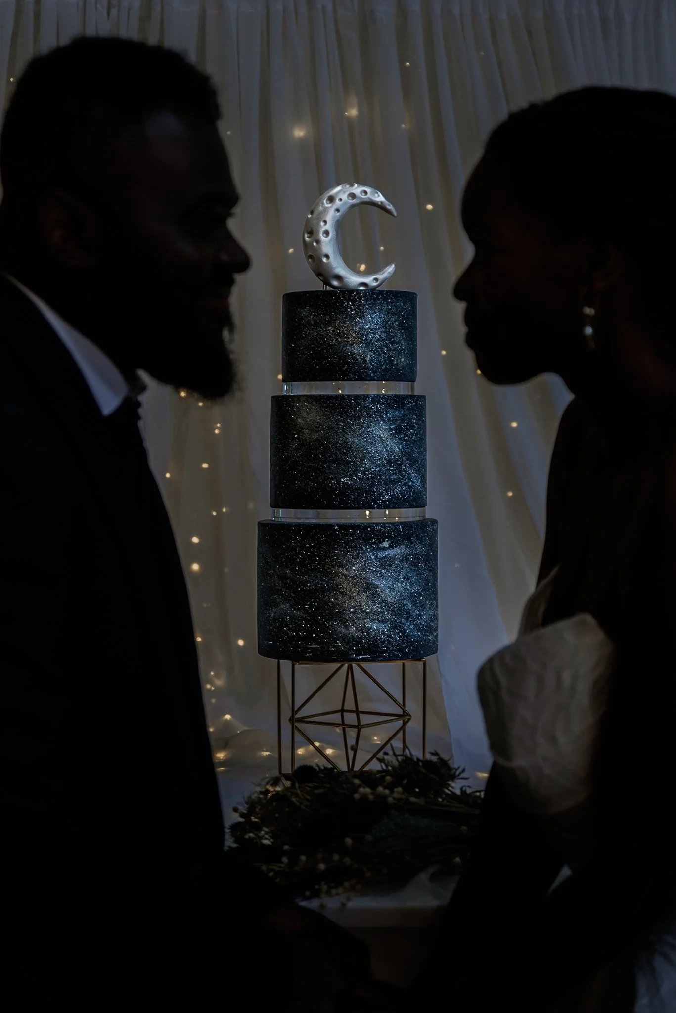 Silhouettes of a man and woman facing each other, with a multi-tiered cake with a galaxy design and a silver moon topper in the background.