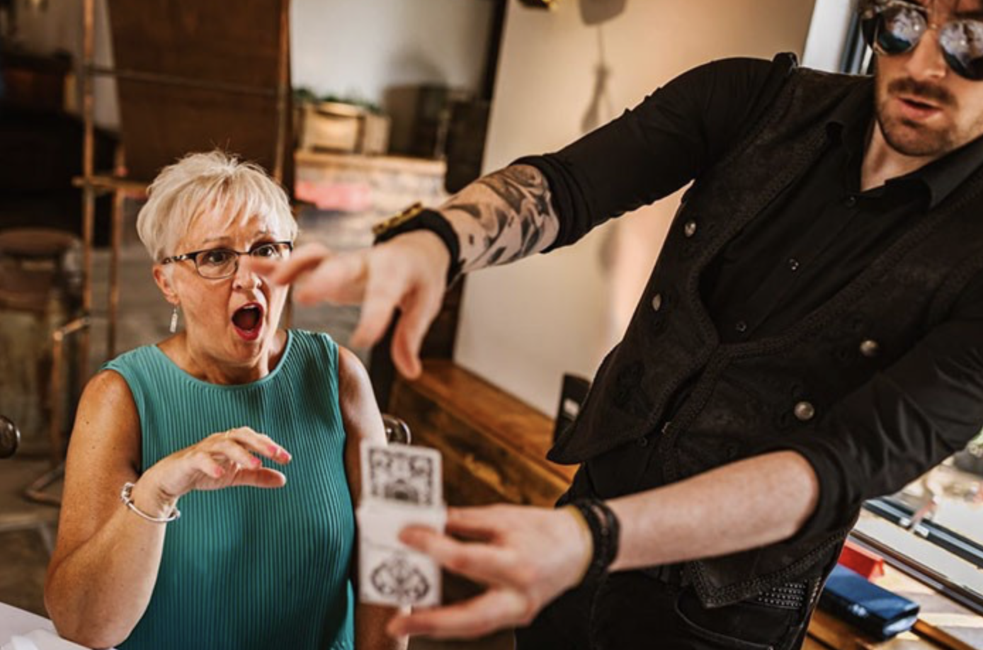 A woman with glasses, short blonde hair, and wearing a sleeveless teal top, looks shocked as a man with tattoos, dark sunglasses, and black clothing performs a magic trick with a card.