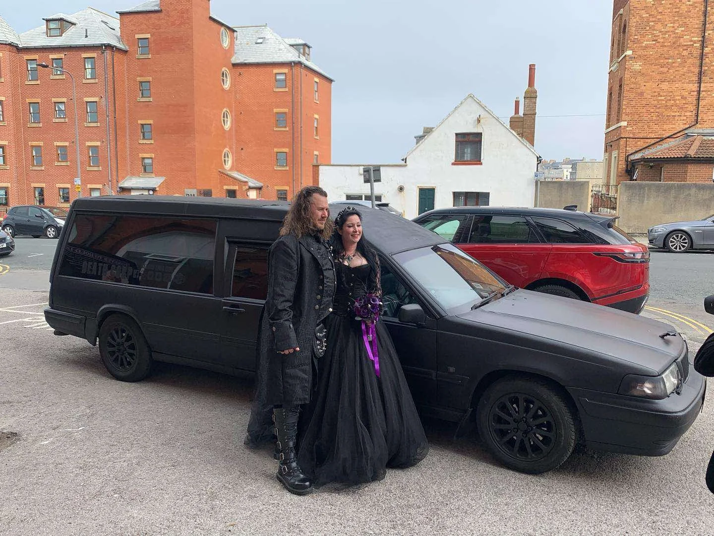 A man and woman in gothic attire standing next to a black station wagon, with the woman holding a bouquet of purple flowers. They are outdoors in a parking lot with brick and white buildings in the background.