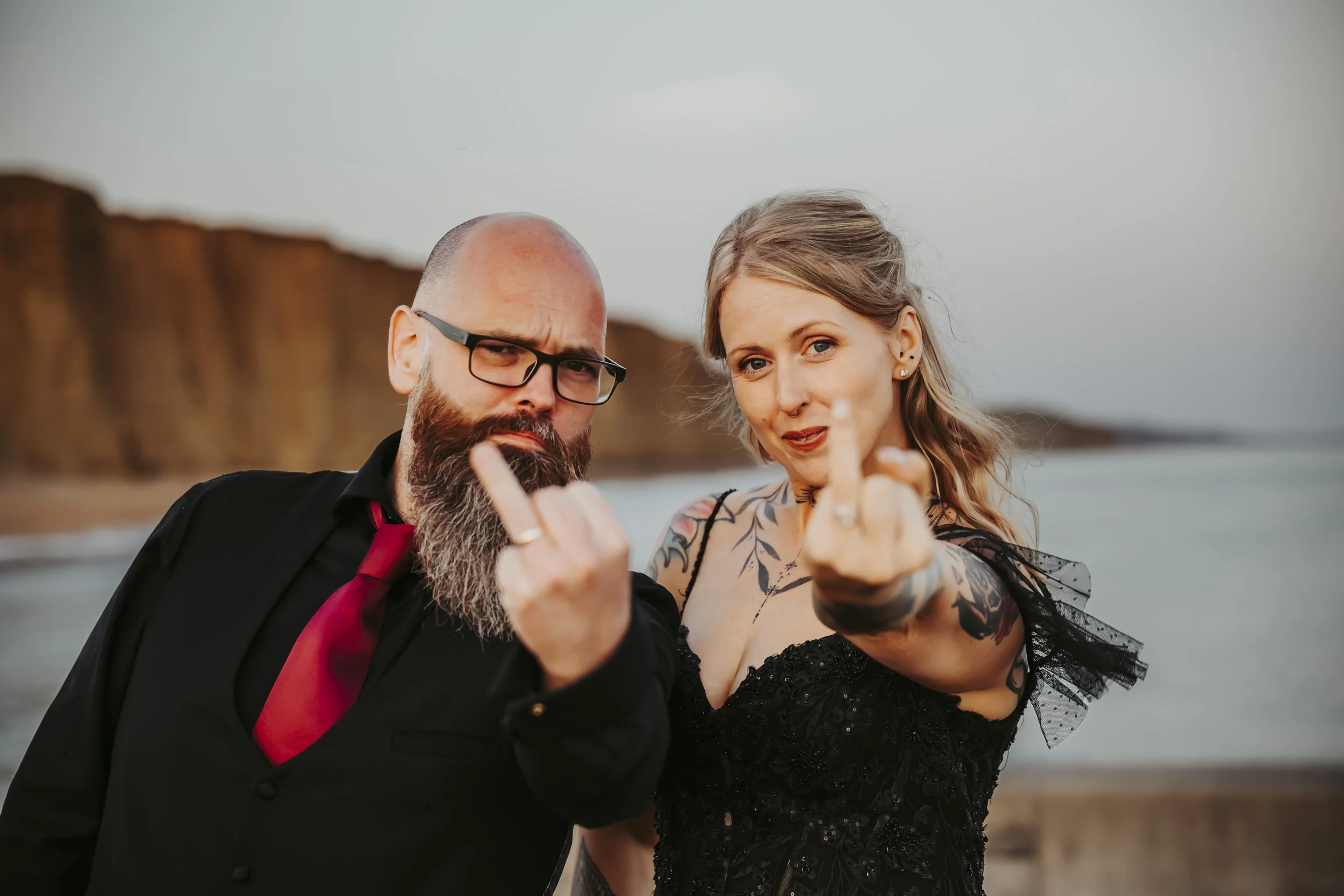 Couple dressed in black wedding outfits hold up their ring fingers towards the camera