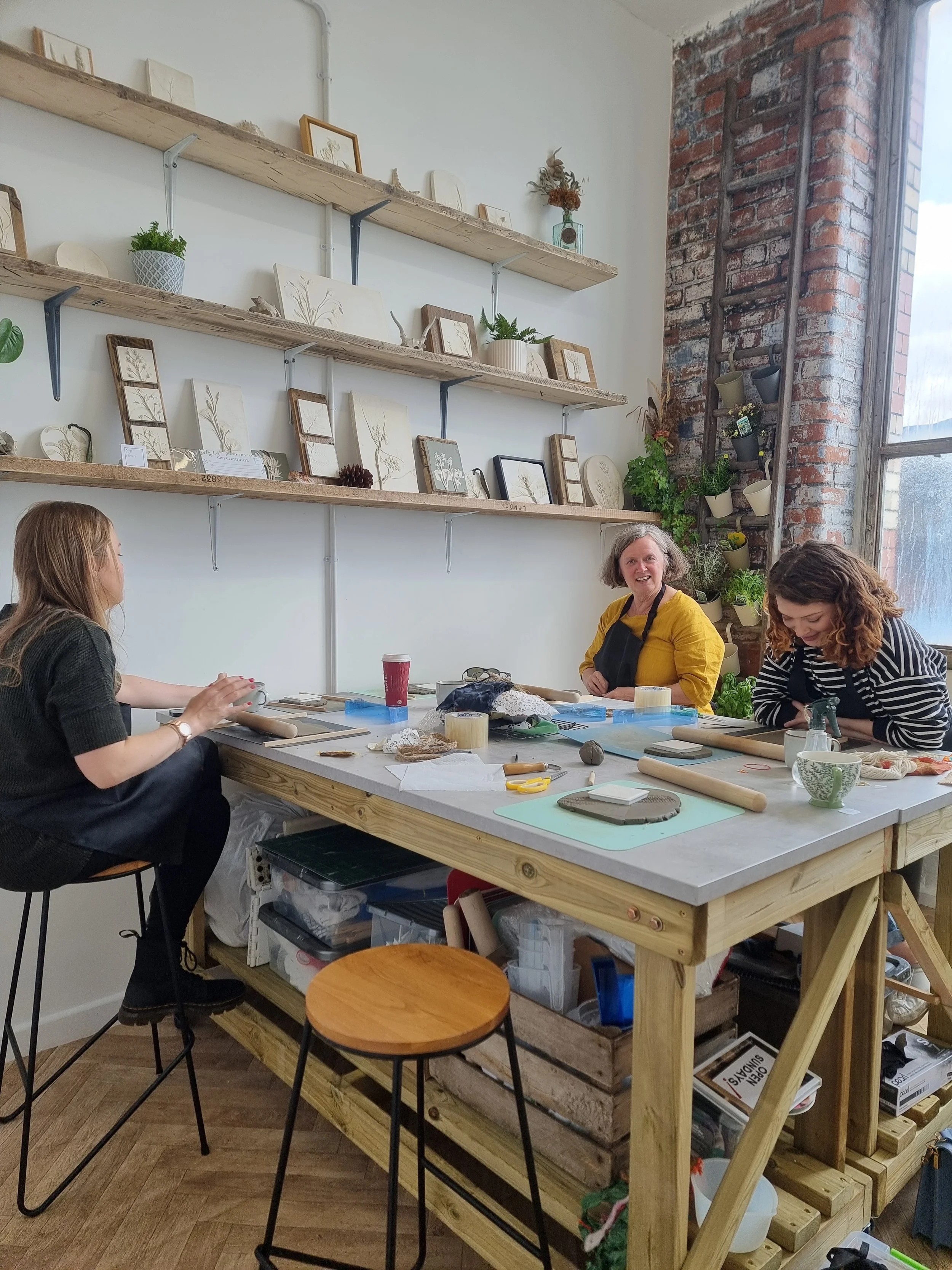 Three women sitting around a wooden table in a ceramics studio, working on pottery projects with various tools and clay. Shelves on the wall display framed botanical sketches, potted plants, and decorative items, with an exposed brick wall and large 