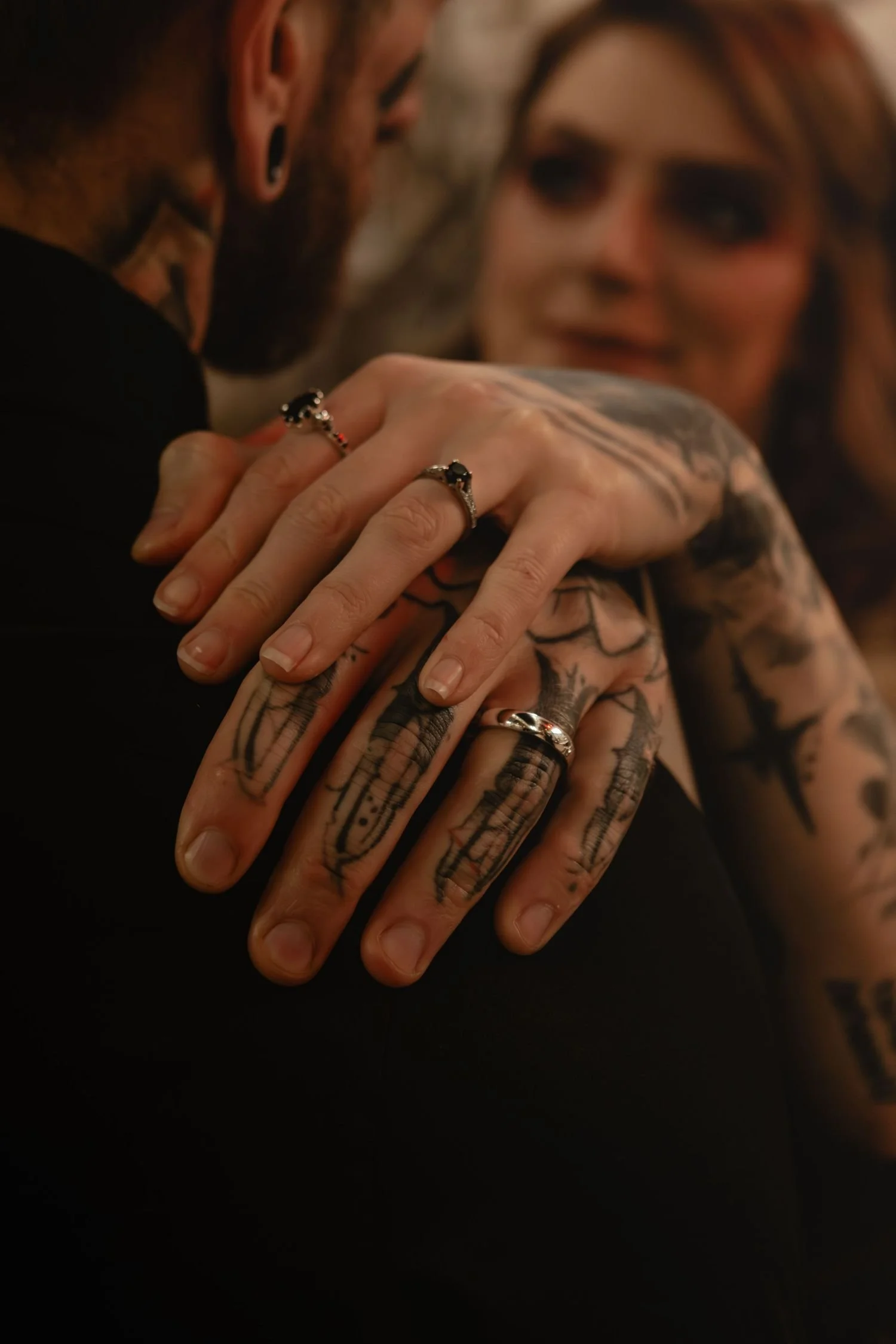 Close-up of a person with multiple tattoos and rings on their fingers, gently holding another person's hand with tattoos, in an intimate moment.