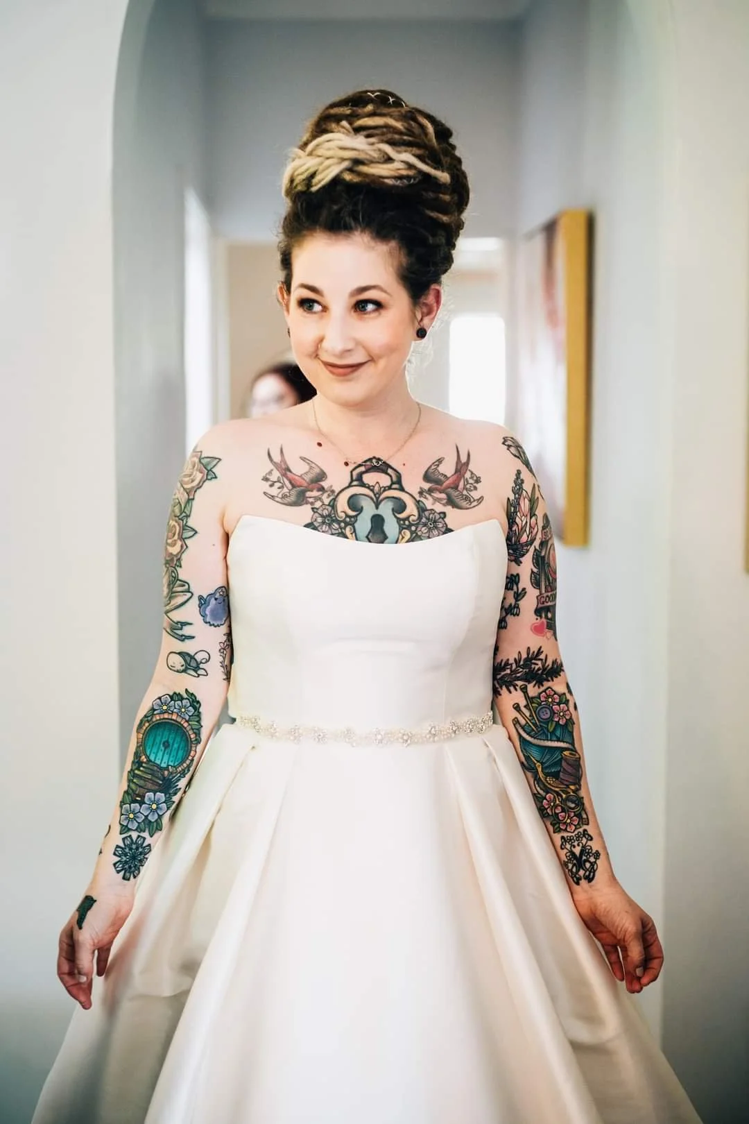 A woman in a white wedding dress with tattoos on her arms and chest, standing in a hallway with a smile.