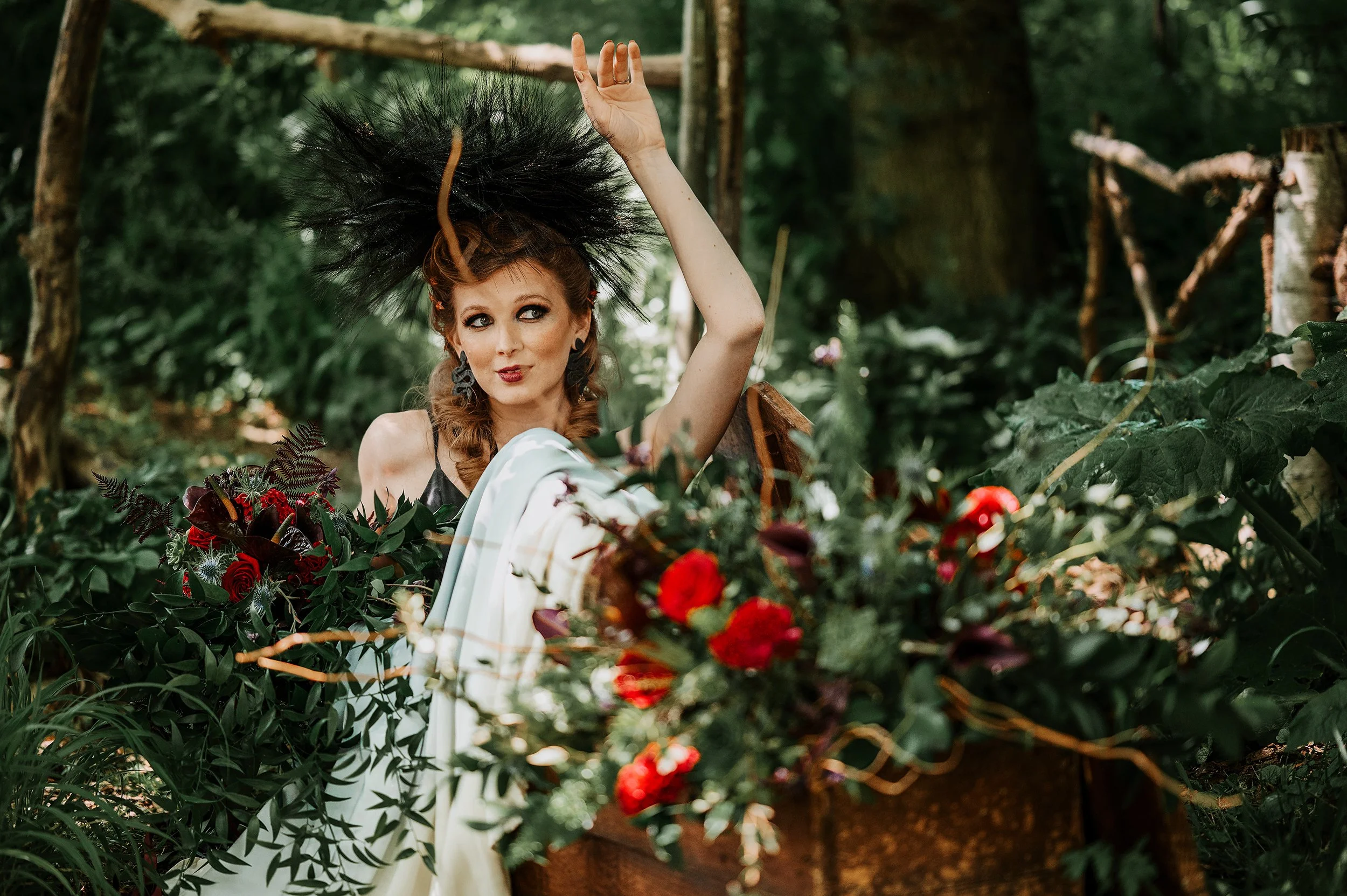 A woman with red hair, wearing a black feathered headpiece and black earrings, is posing in a lush green forest. She is standing behind a large wooden basket filled with red and purple flowers and greenery, with her right hand raised above her head a