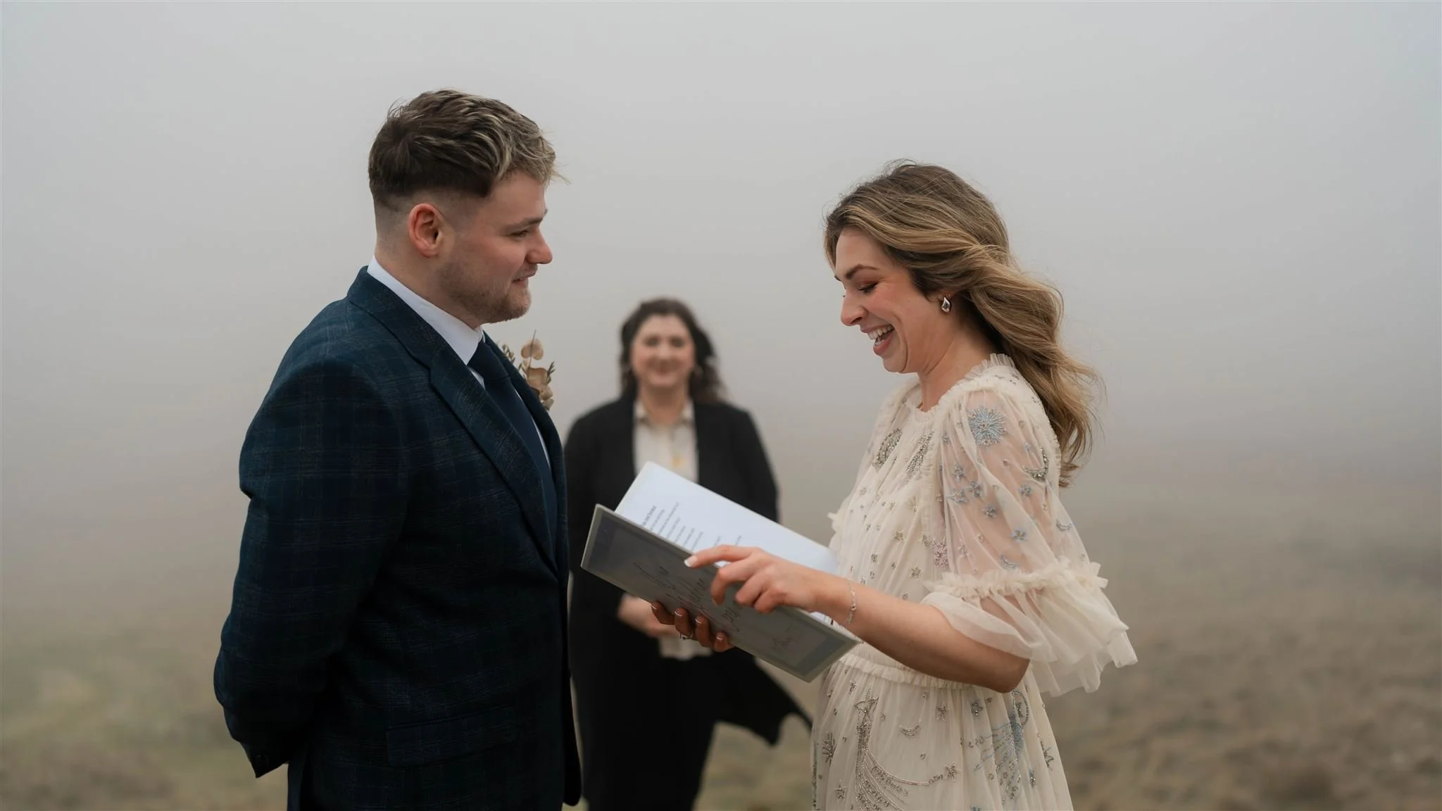 A woman in a light-colored, floral dress is smiling and reading vows from a booklet to a man in a dark plaid suit during an outdoor wedding ceremony on a foggy day. Another woman in black is smiling in the background.