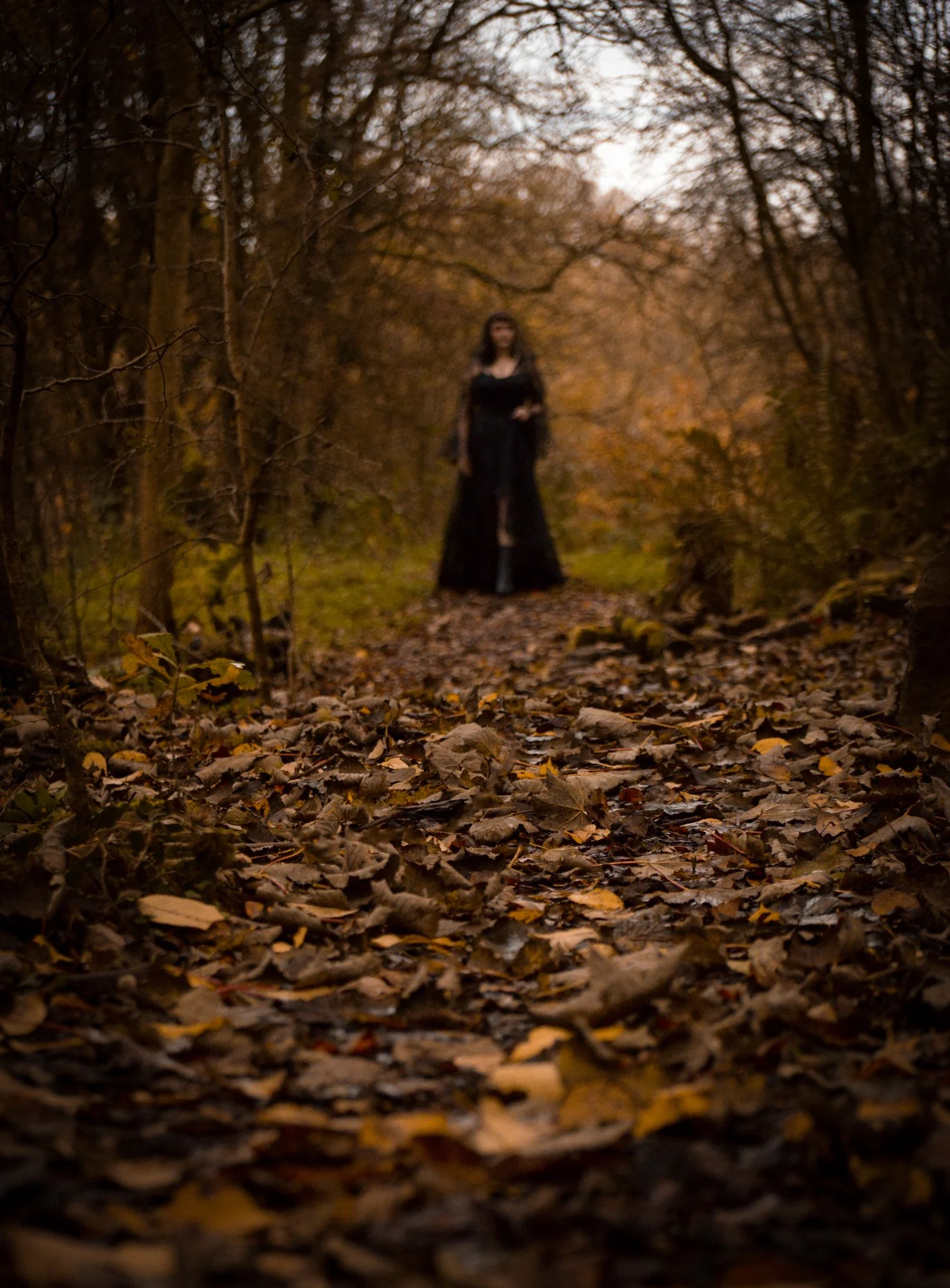 A woman in a long black dress standing on a leaf-covered forest trail during autumn, with blurred trees in the background.