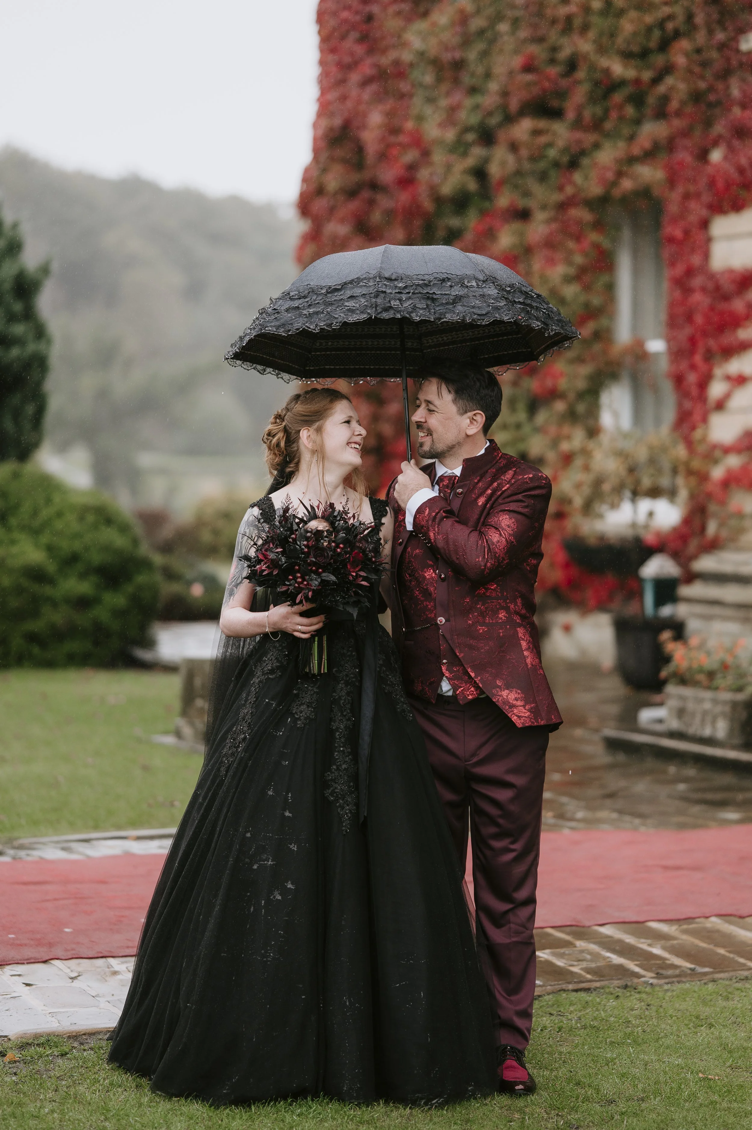 A couple in formal attire on their wedding day standing under a black umbrella in the rain, smiling at each other, with a red carpet on the ground and a backdrop of a house covered in red and green foliage.