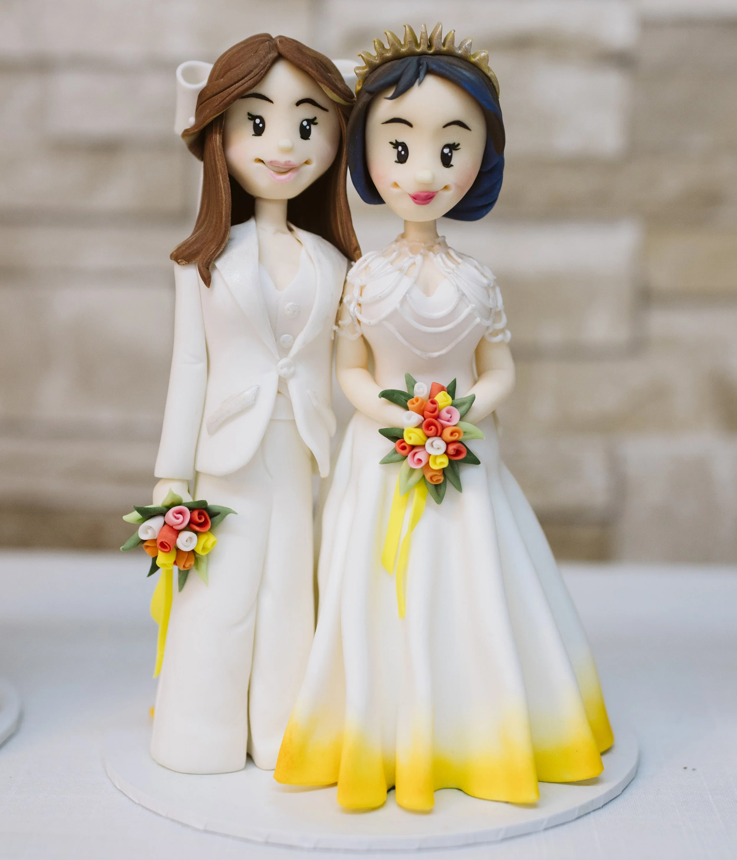 Cake topper of two women in wedding attire, one in a white suit and the other in a white wedding dress with yellow accents, both holding small bouquets, against a neutral background.