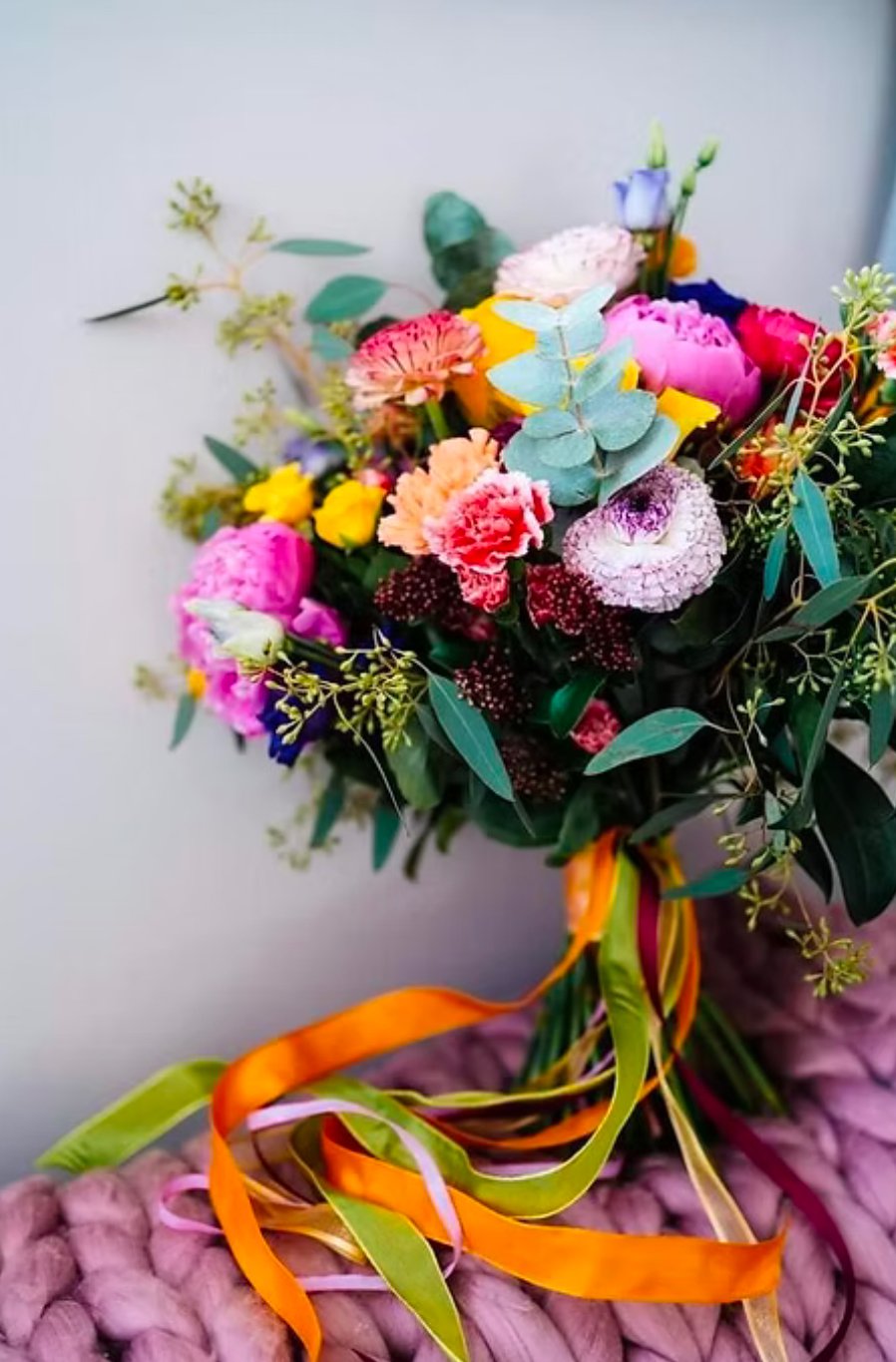 Colorful bouquet of various flowers including pink, yellow, purple, and white, tied with multicolored ribbons, resting on a pink knitted surface.