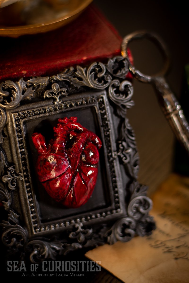 A decorative framed model of a human heart, colored in red, leaning against intricate black gothic-style frame with scrollwork. A small part of a key is visible to the right, and a piece of parchment with writing is partially seen at the bottom.