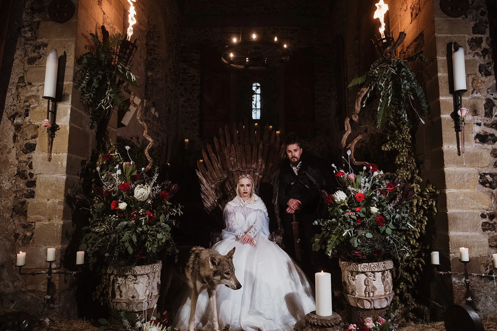 A woman in a white wedding gown and a man in dark clothing sitting in front of a throne-like chair, surrounded by large floral arrangements, candles, and torches, with a wolf walking in front.