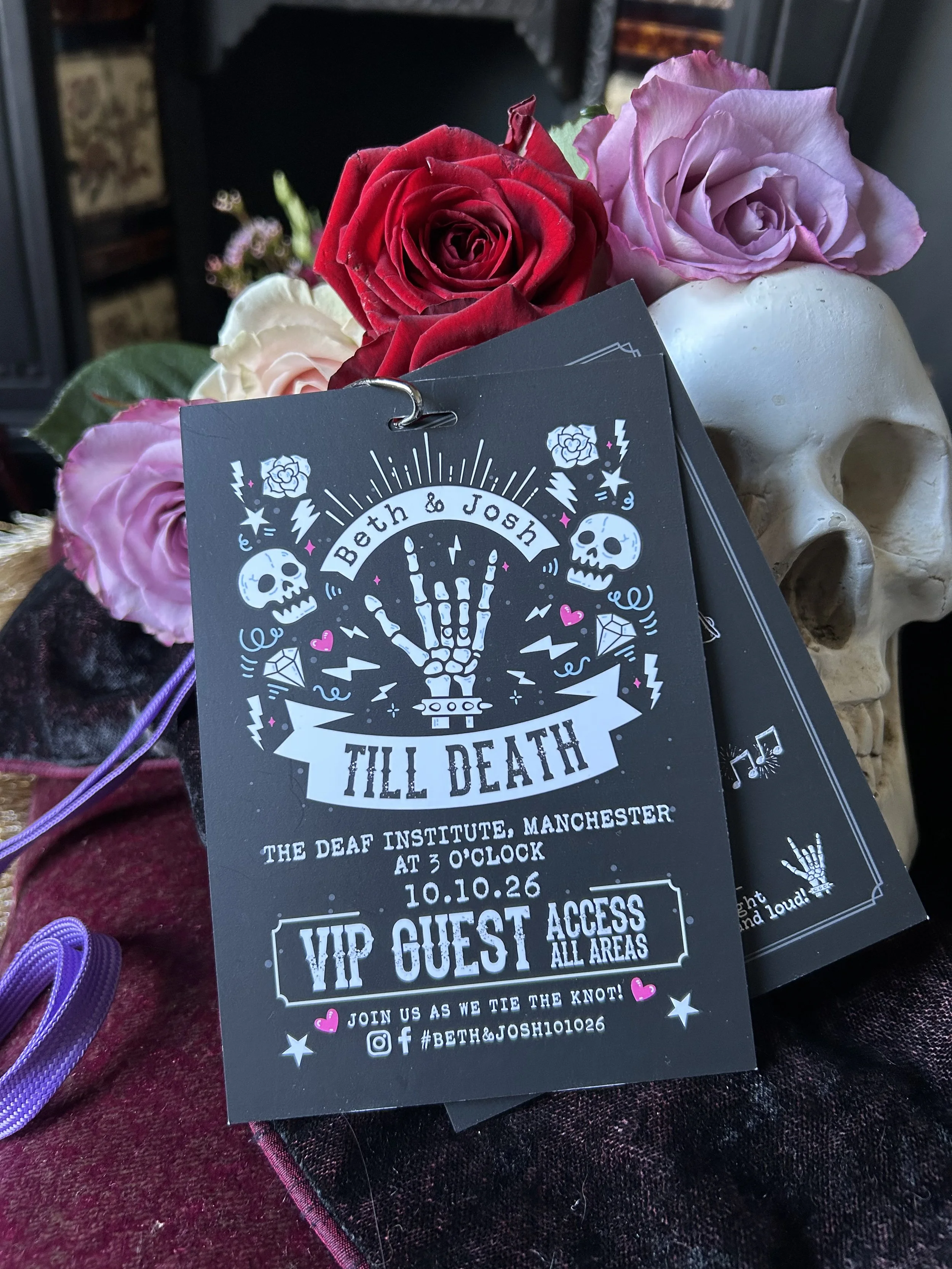 A wedding invitation for Beth and Josh featuring gothic-style artwork, including a hand skeleton, skulls, and hearts, placed among pink and red roses with a white skull decoration in the background.