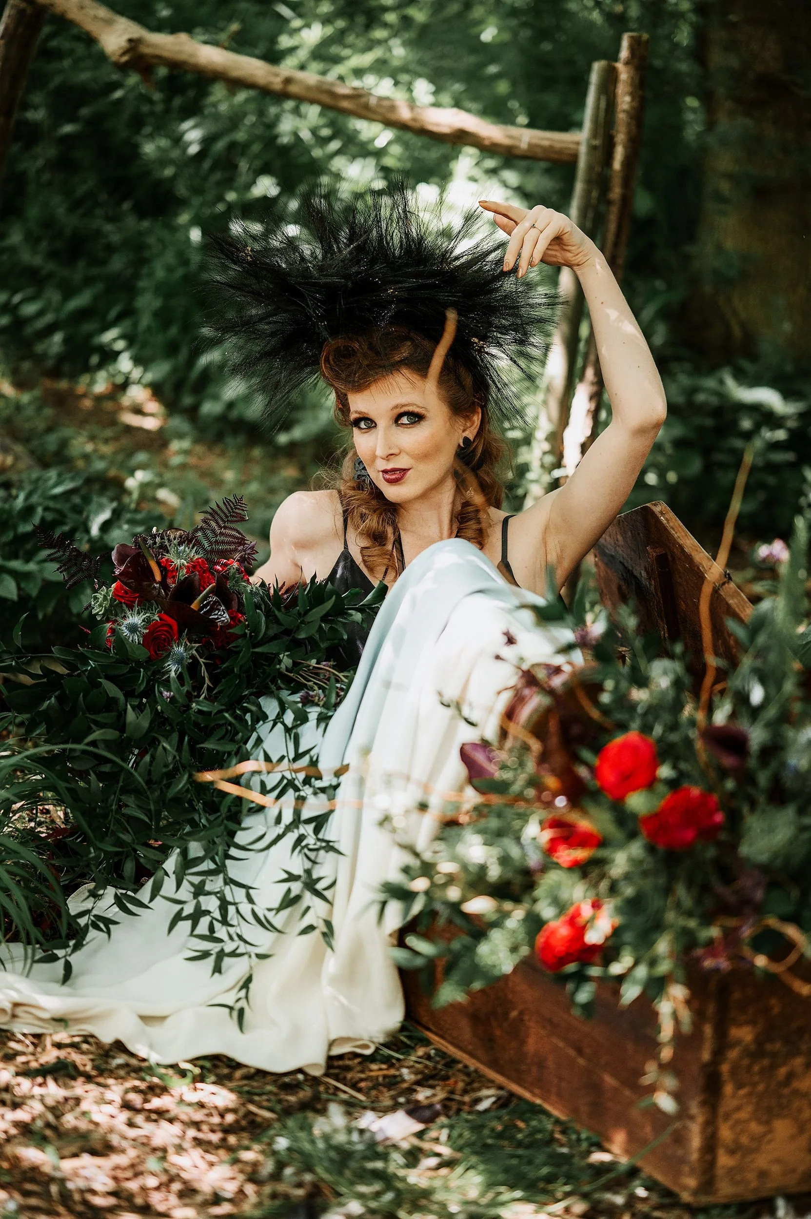 A woman with elaborate black feathered headpiece, makeup, and vintage style dress sitting amidst greenery and flowers in a wooded outdoor setting.