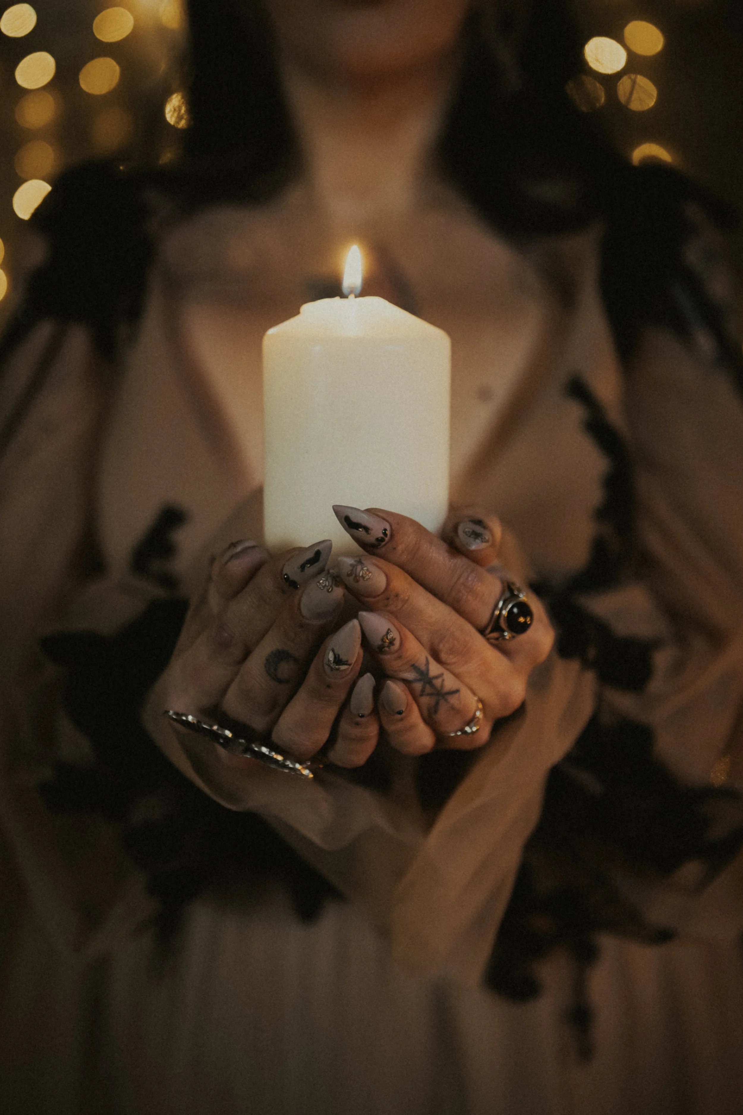 Person holding a lit white candle with patterned manicured nails and rings, with a dark background