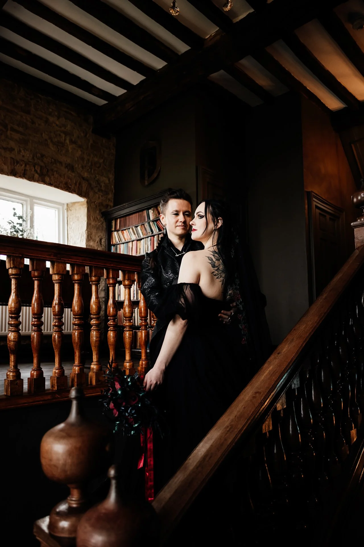 Two women standing close on a staircase inside a rustic house, with dark hair, tattoos, and gothic style clothing, one holding a bouquet of flowers.