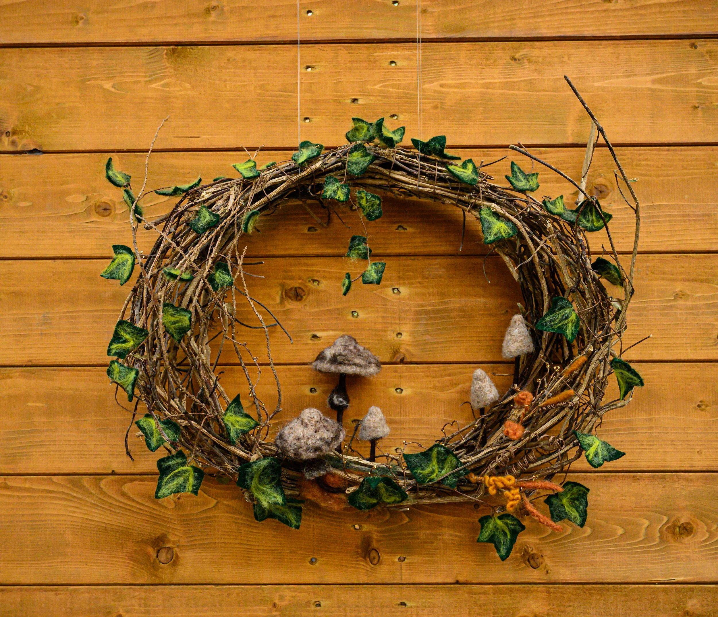 A decorative wreath made of intertwined twigs and vines, decorated with green ivy leaves and felt mushrooms, hanging against a wooden wall.