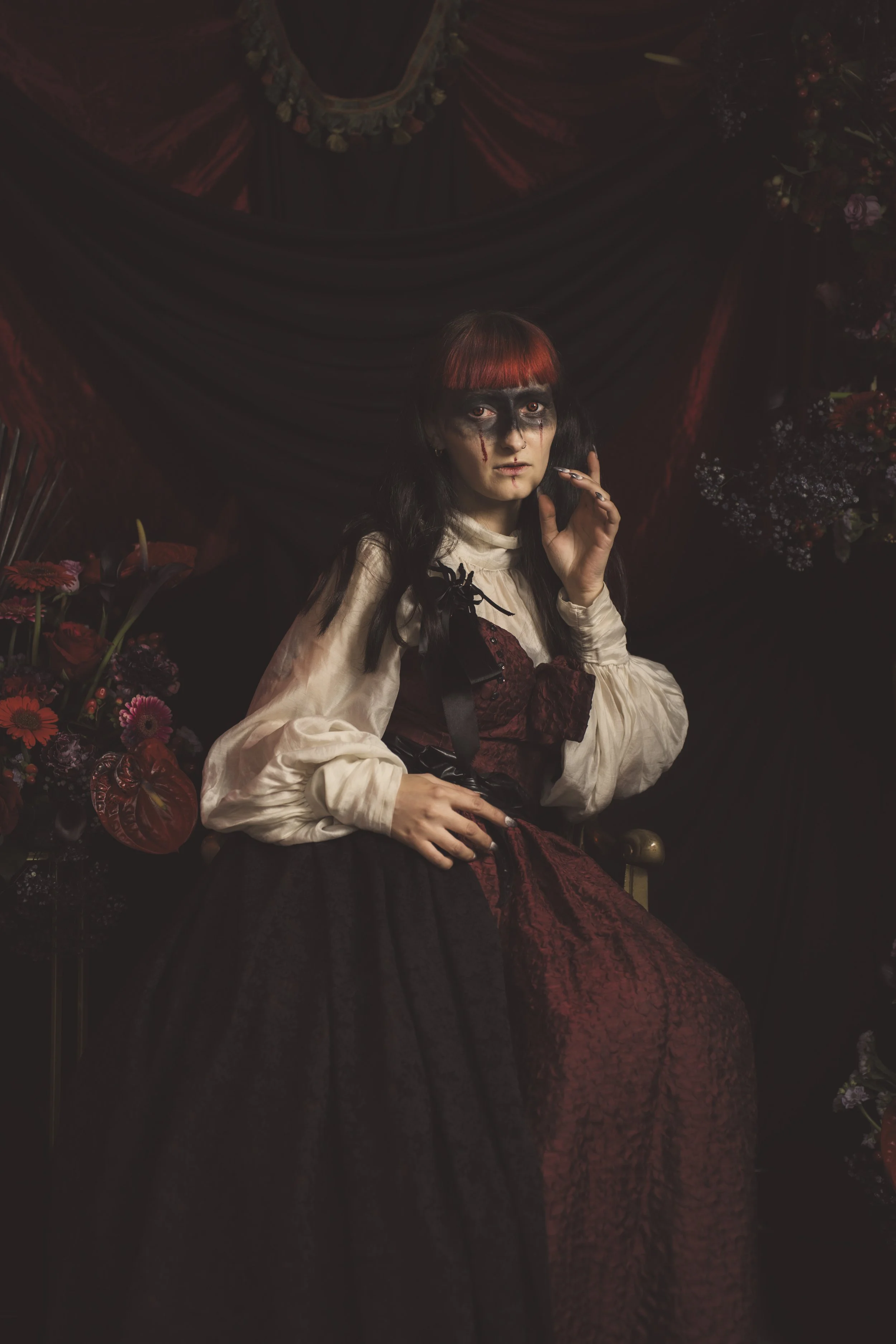 A young woman with black and red hair, dark eye makeup, and face paint sitting on a vintage chair against a dark, floral backdrop. She wears a long, dark, Victorian-style dress with a cream blouse and is surrounded by dark flowers.