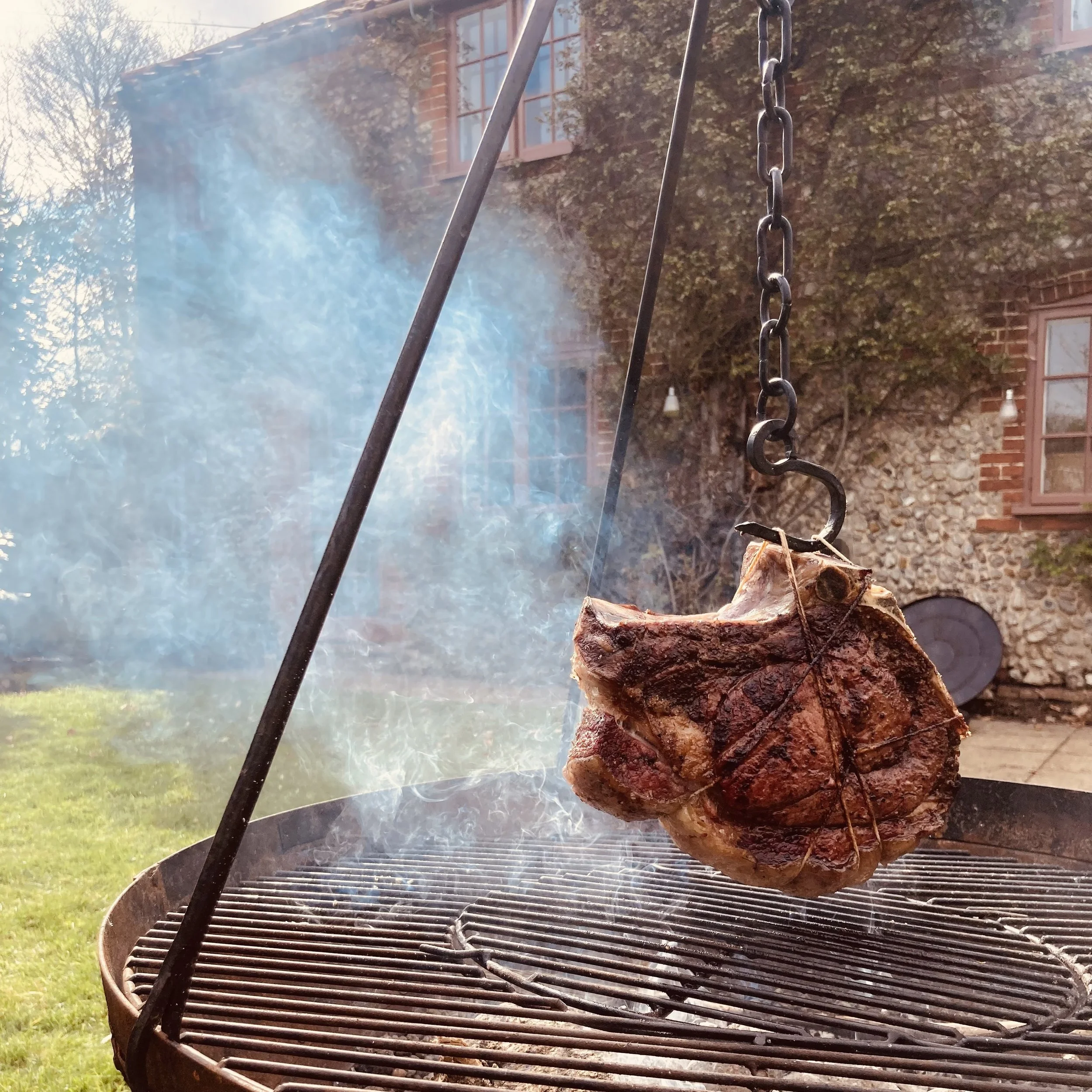 A large piece of meat hanging over a round grill outdoors, with smoke and a brick house with windows in the background.
