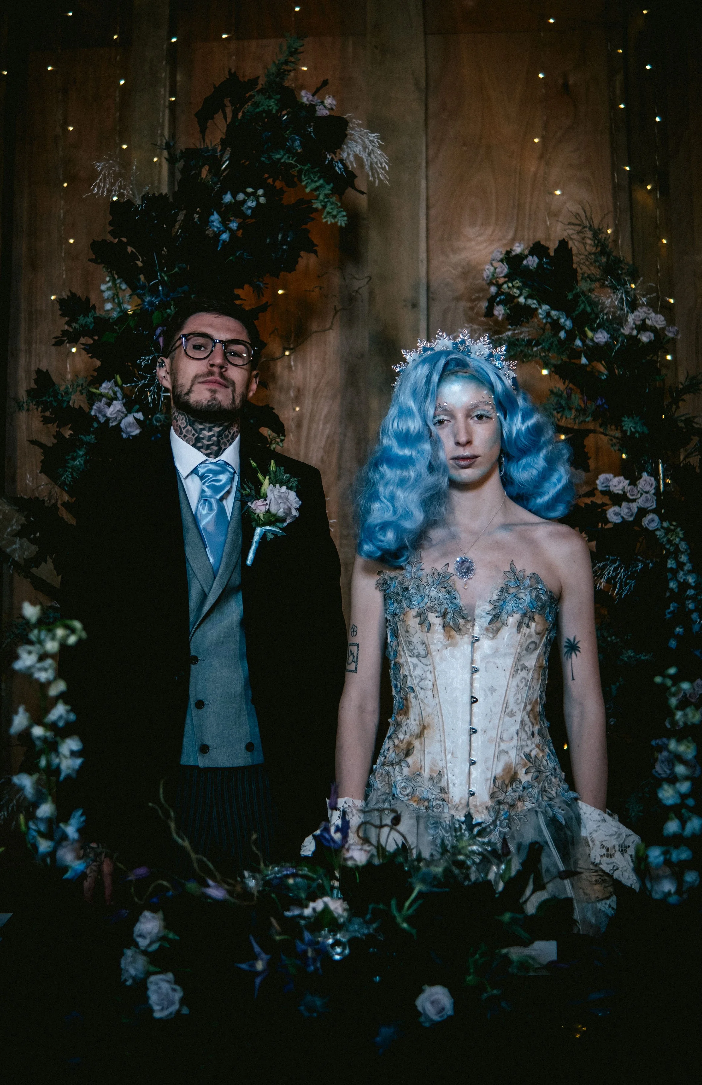 A couple dressed in wedding attire stand in front of a floral arrangement with a wooden backdrop. The man has dark hair, glasses, and tattoos, wearing a black suit with a gray vest and light blue tie. The woman has vibrant blue, curly hair, and a tat