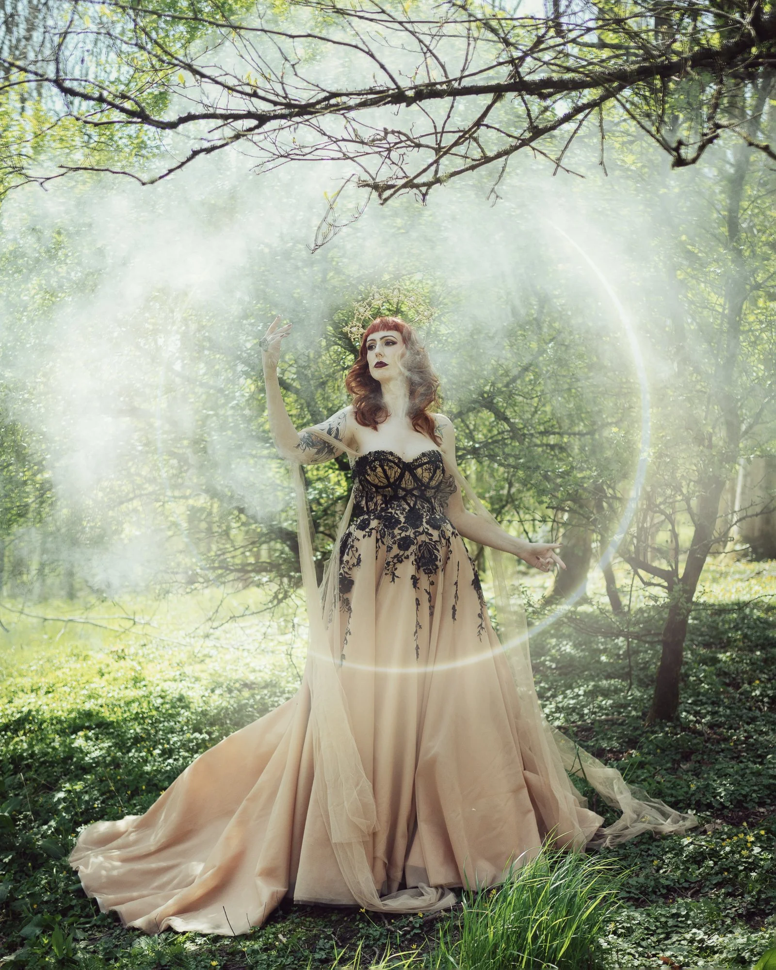 A woman in a beige and black lace gown standing in a forest with sunlight shining through trees, surrounded by mist and a glowing light orb.