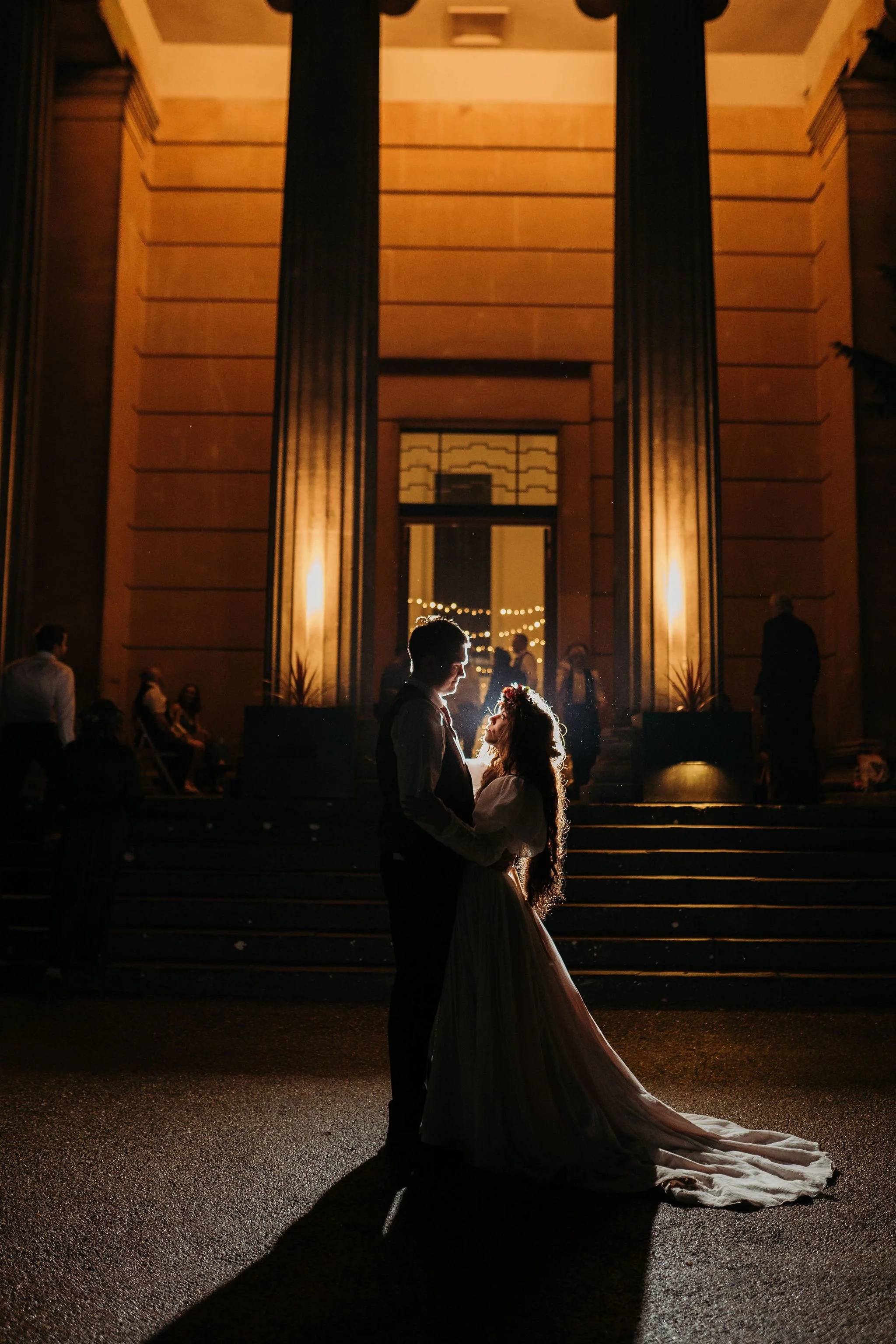 Silhouette of a bride and groom embracing at night in front of a large illuminated building with steps, surrounded by a few guests.