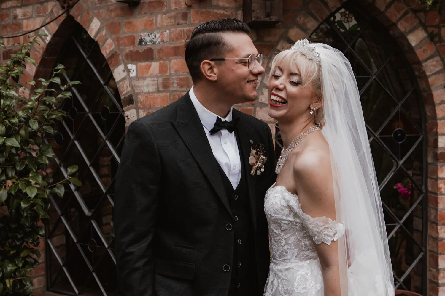 A smiling bride and groom in wedding attire share a joyful moment outdoors near a brick wall with arched windows.