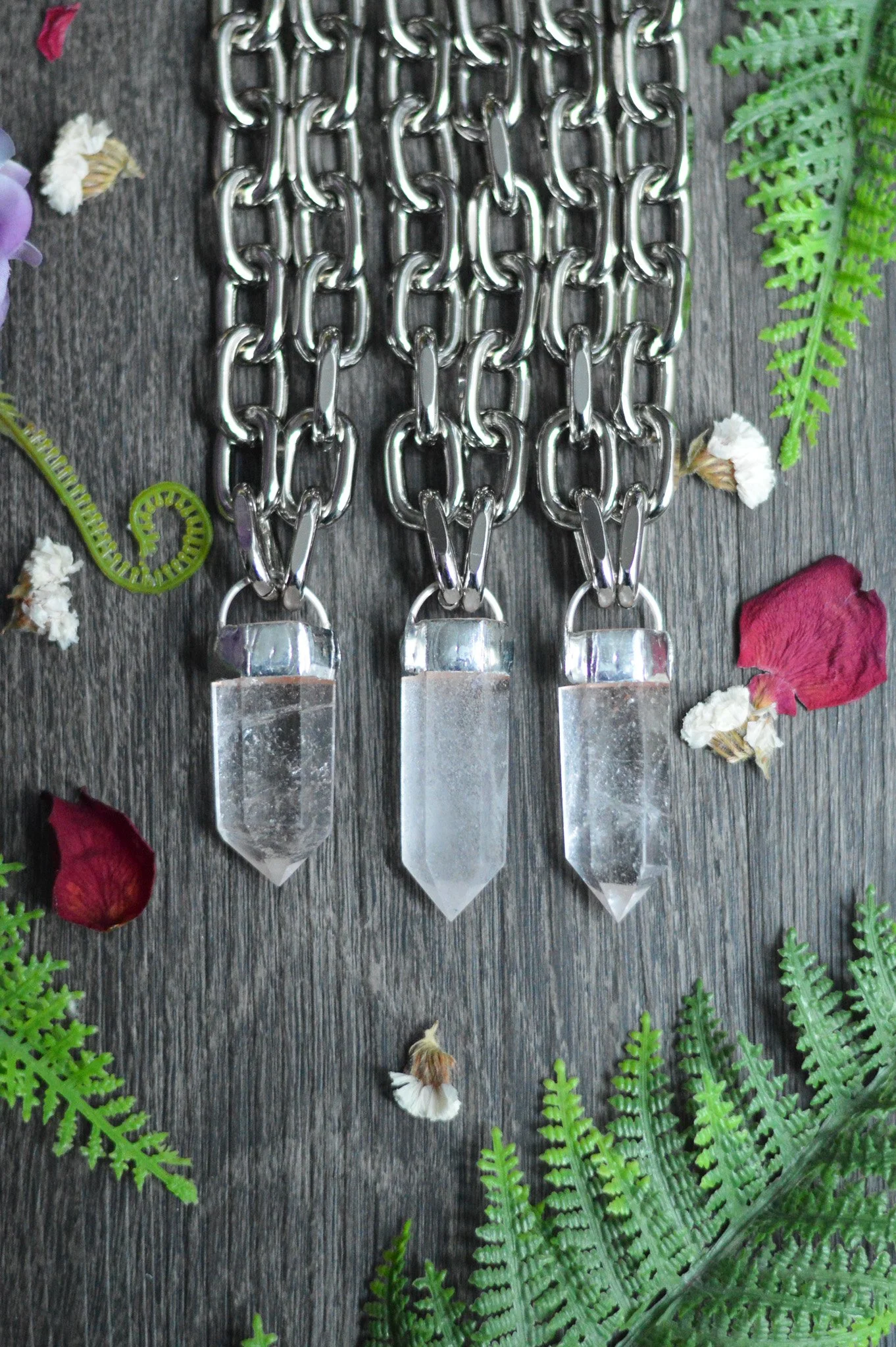 Three crystal prism pendants hanging from silver chain links, surrounded by fern leaves and flower petals on a wooden surface.