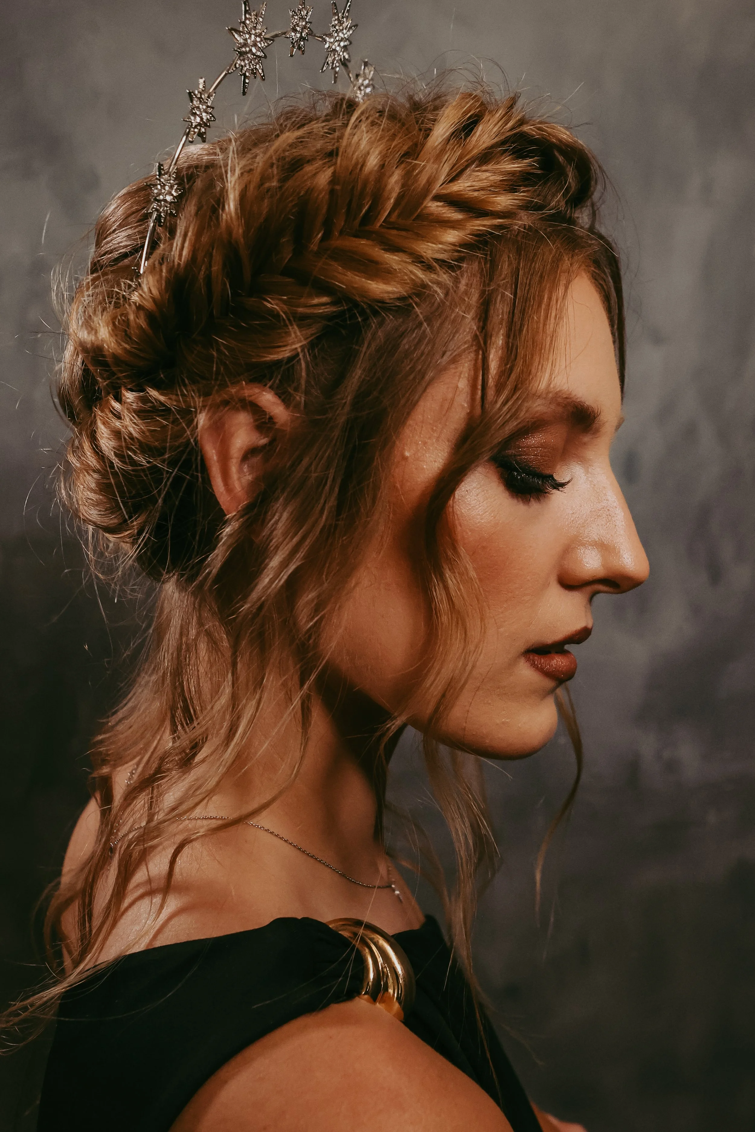 A woman with wavy, reddish-brown hair styled in an updo, wearing a star-shaped headband and a black dress with gold embellishments on the shoulder, against a dark gray background.