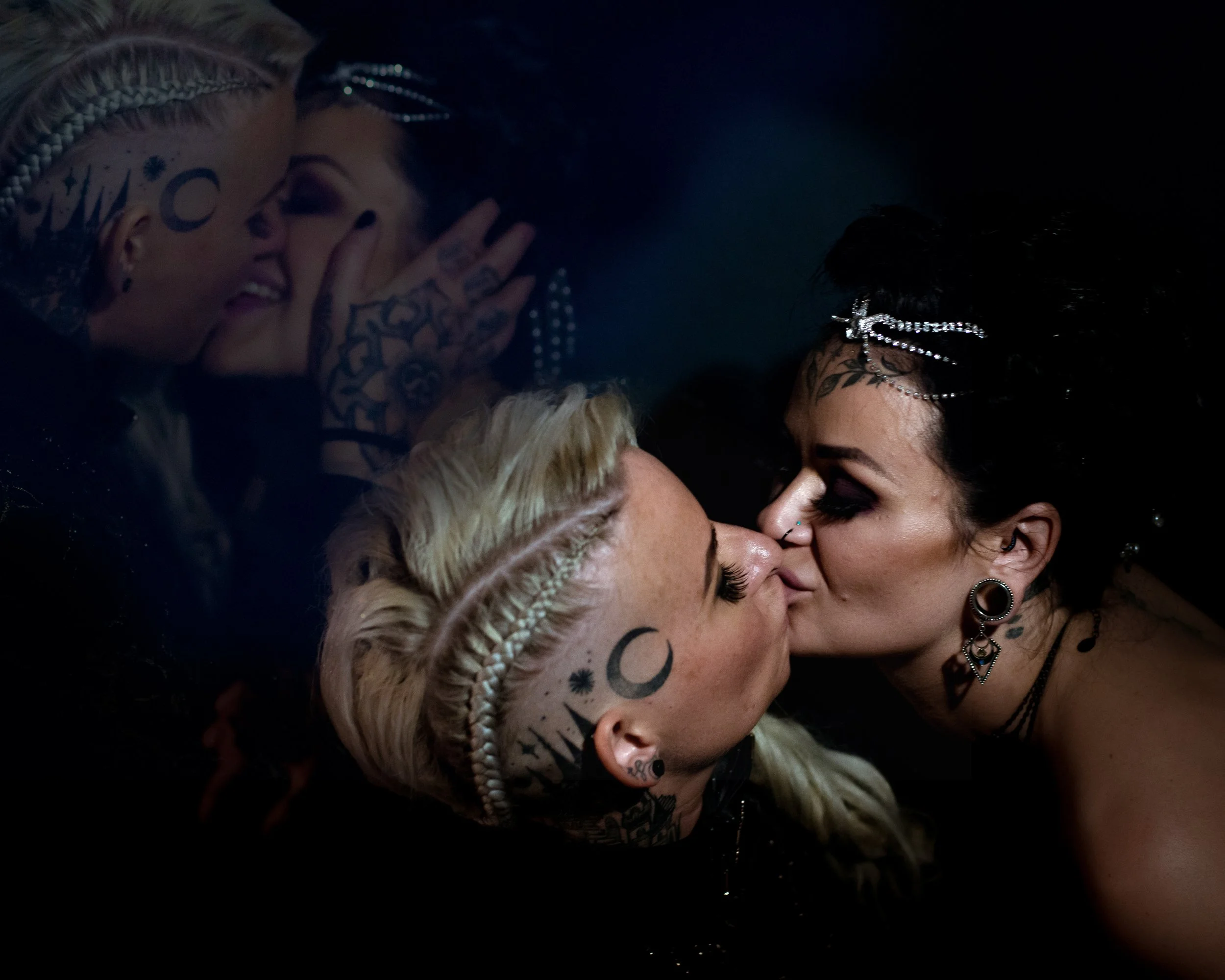 Two women with tattoos and piercings kissing, their reflections visible in water or a mirror.