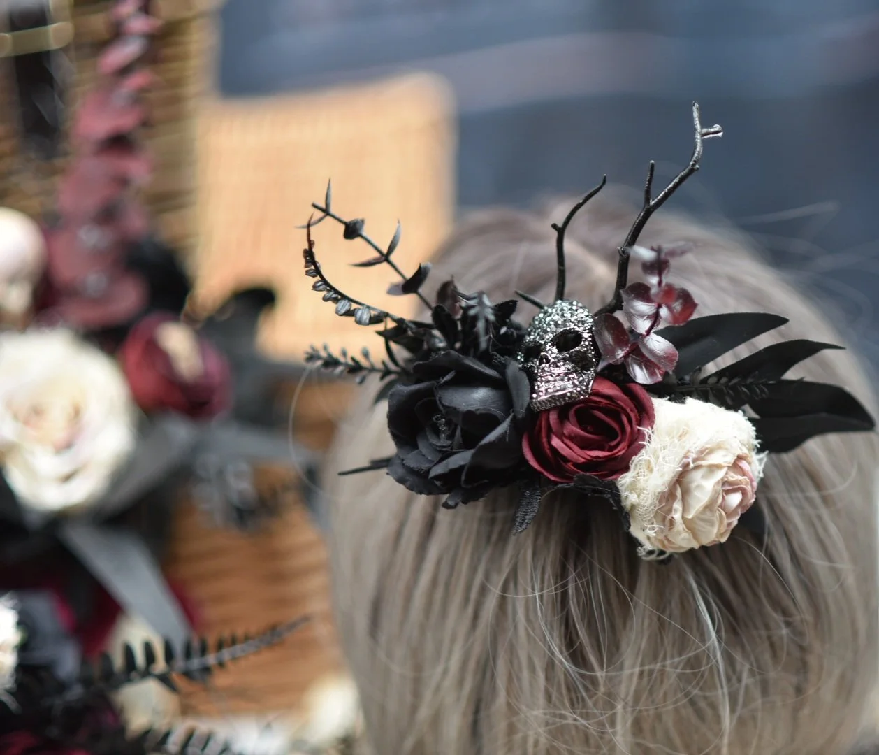 A headband with gothic and floral decorations including a silver skull, roses, black leaves, and branches worn on a person's head with blonde hair.
